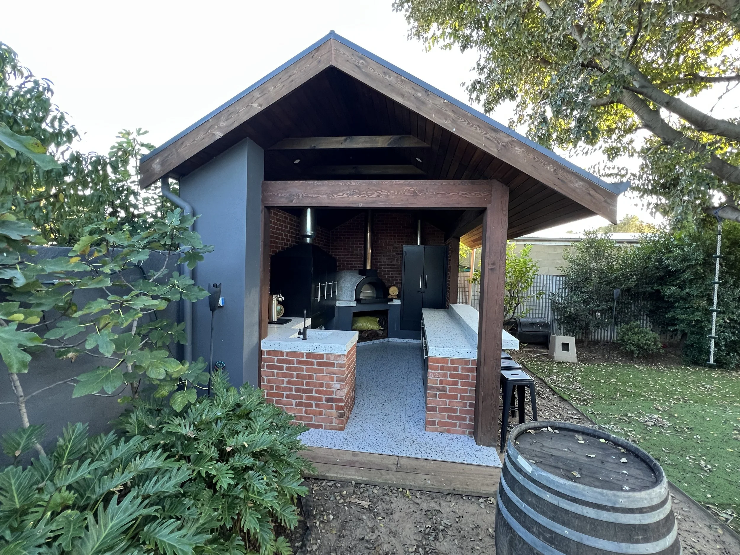 Covered outdoor kitchen with a wood-fired pizza oven, countertops, cabinets, and brick pillars in a landscaped backyard
