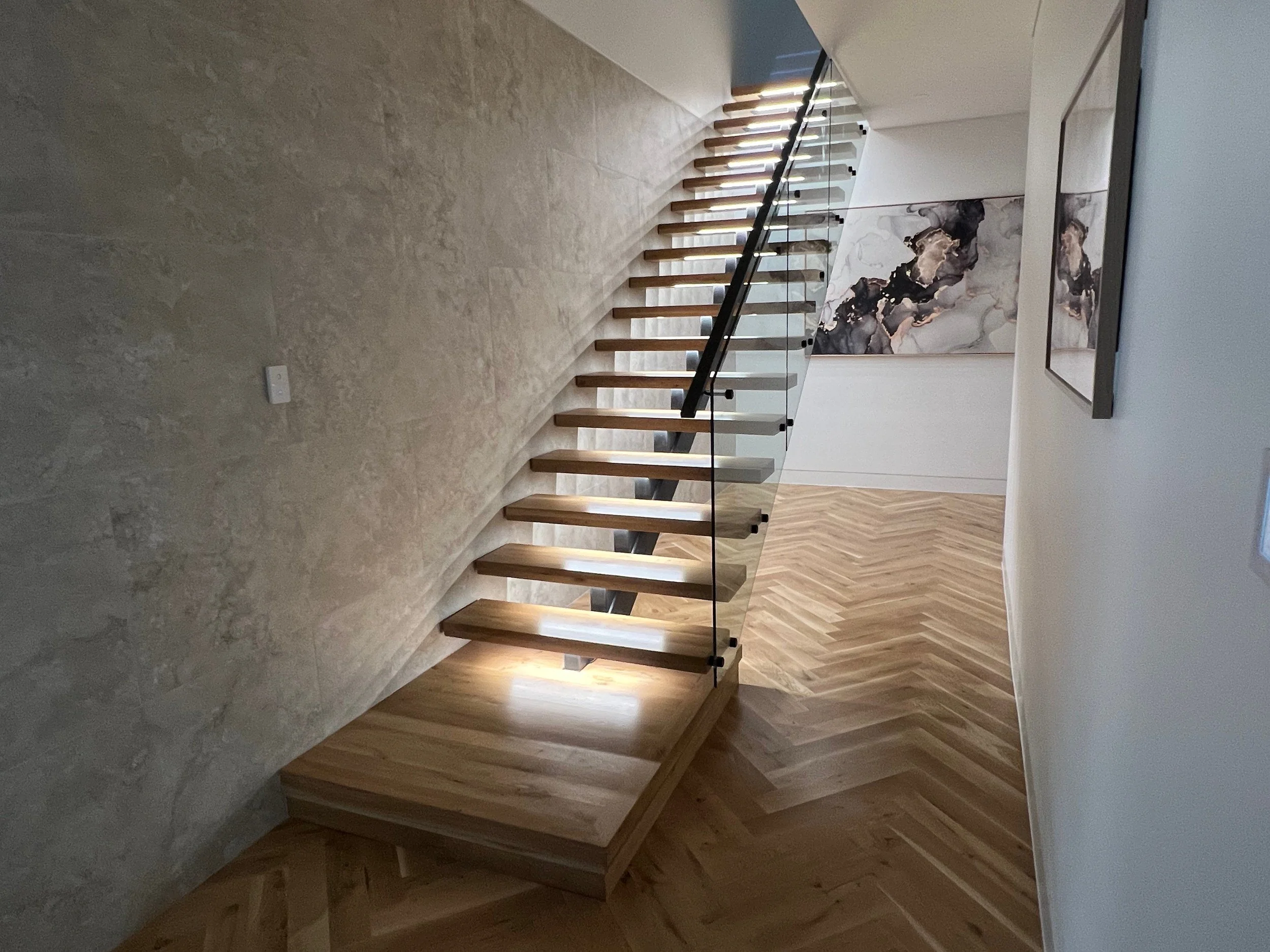 Modern floating wood staircase with glass railing, textured concrete wall, herringbone wood flooring, and abstract wall art