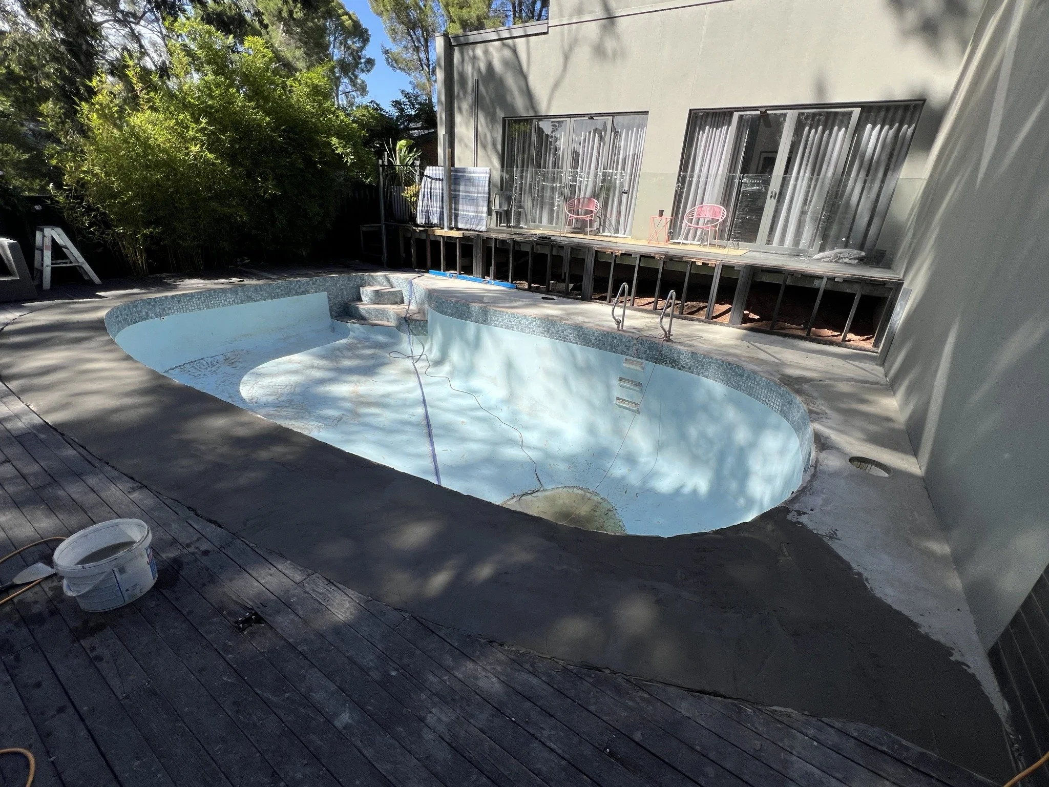 Empty in-ground swimming pool under renovation beside a modern house, with exposed deck supports, patio chairs, trees, and a paint bucket.