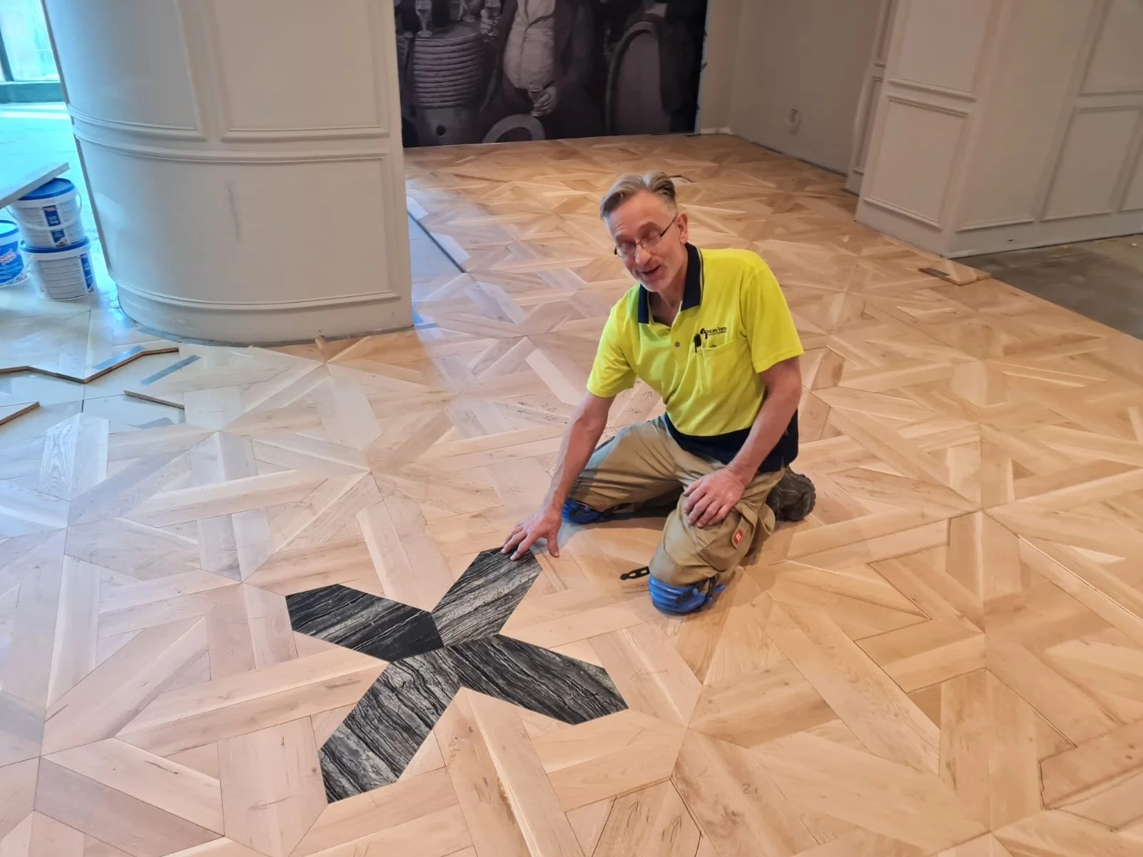 Flooring installer kneeling beside an ornate geometric parquet wood floor with a dark marble inlay