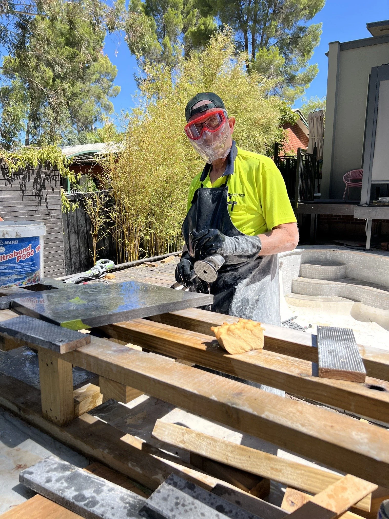 Worker wearing protective goggles, gloves, and apron uses a grinder on stone slabs outdoors.