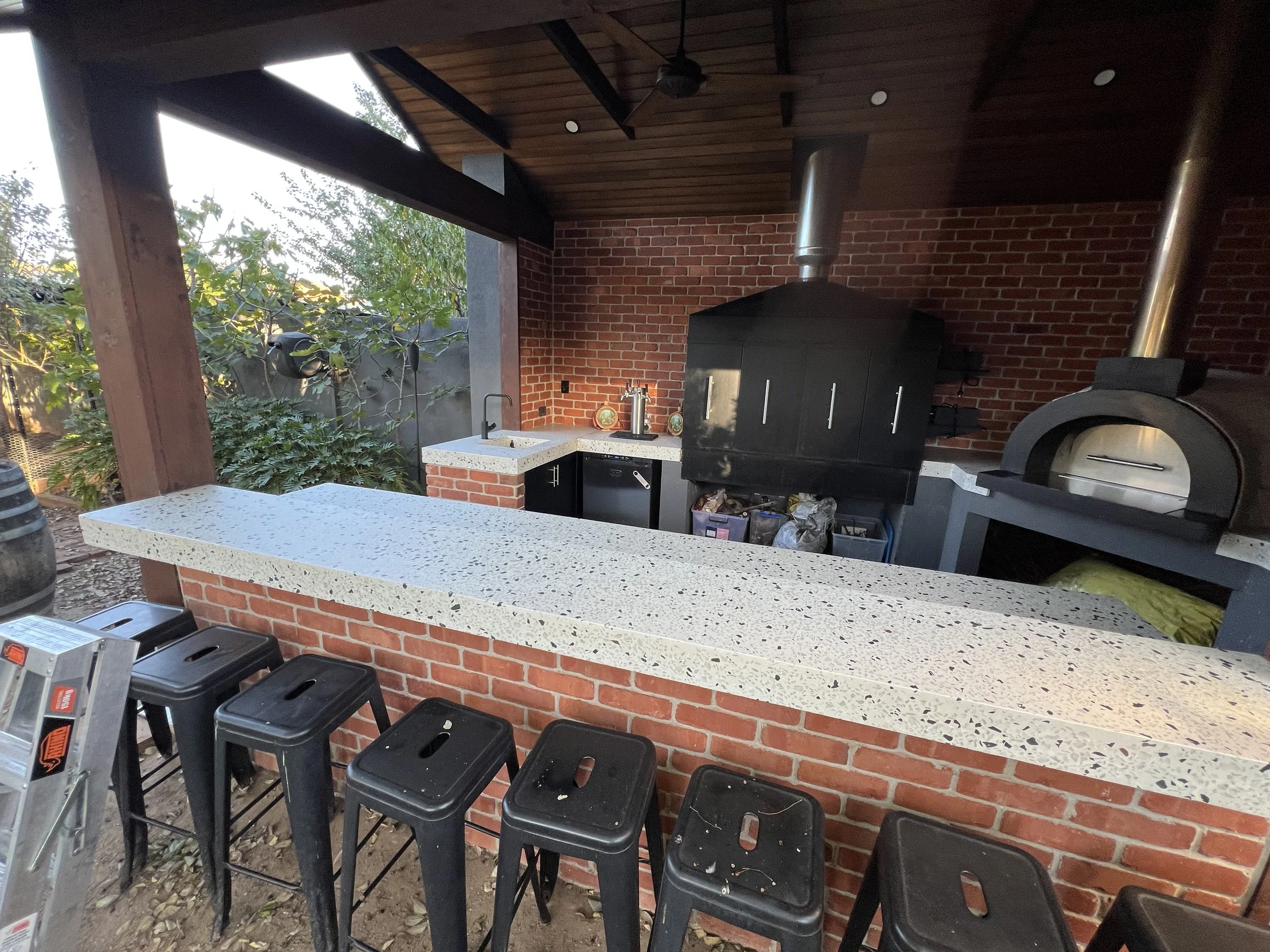 Outdoor covered kitchen with brick walls, terrazzo countertops, black barbecue grill, wood-fired pizza oven, sink, beer taps, and bar stools