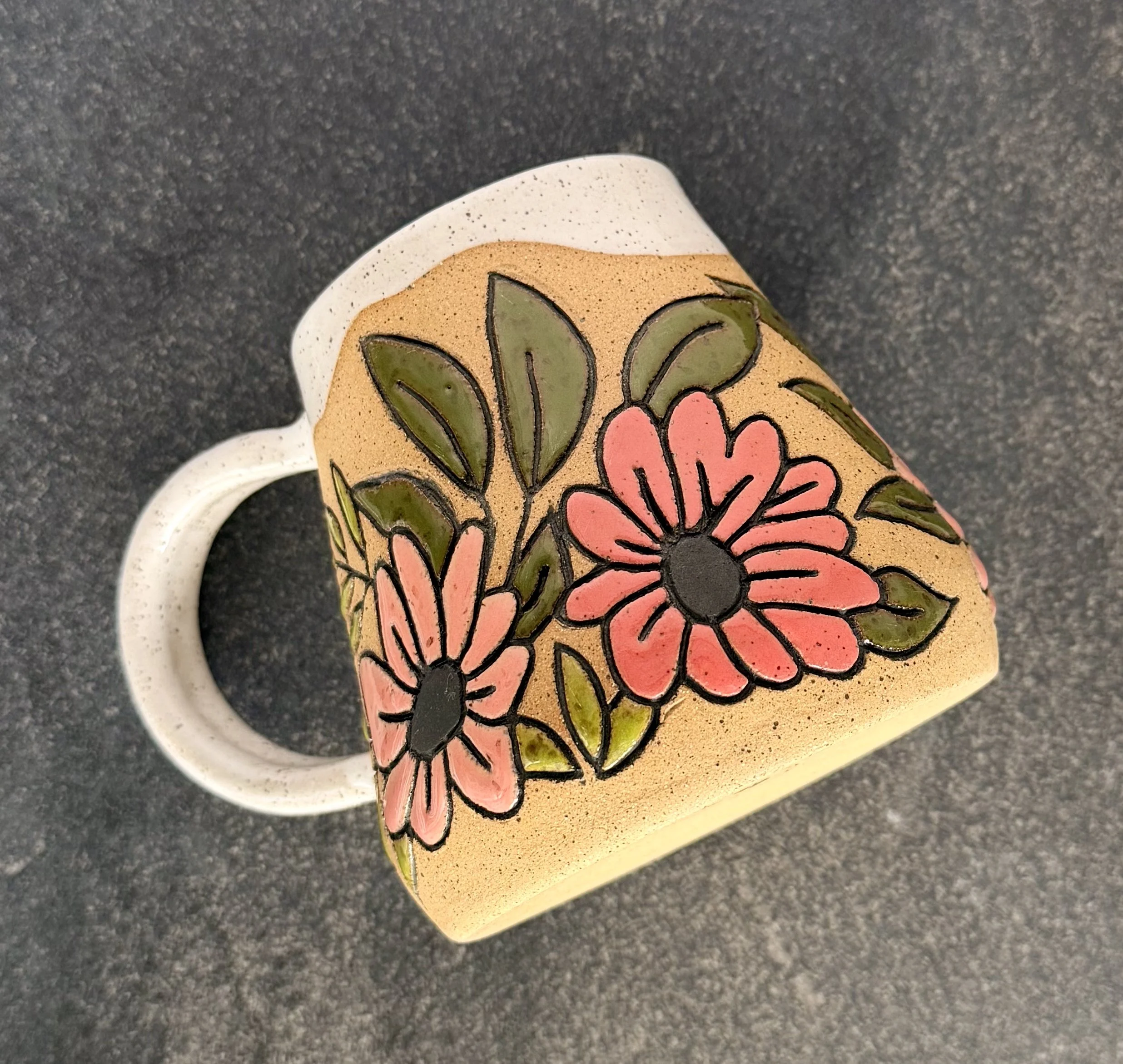 A ceramic mug with floral and leaf designs in pink, green, and black on a raw speckled clay background, placed on a dark gray textured surface.