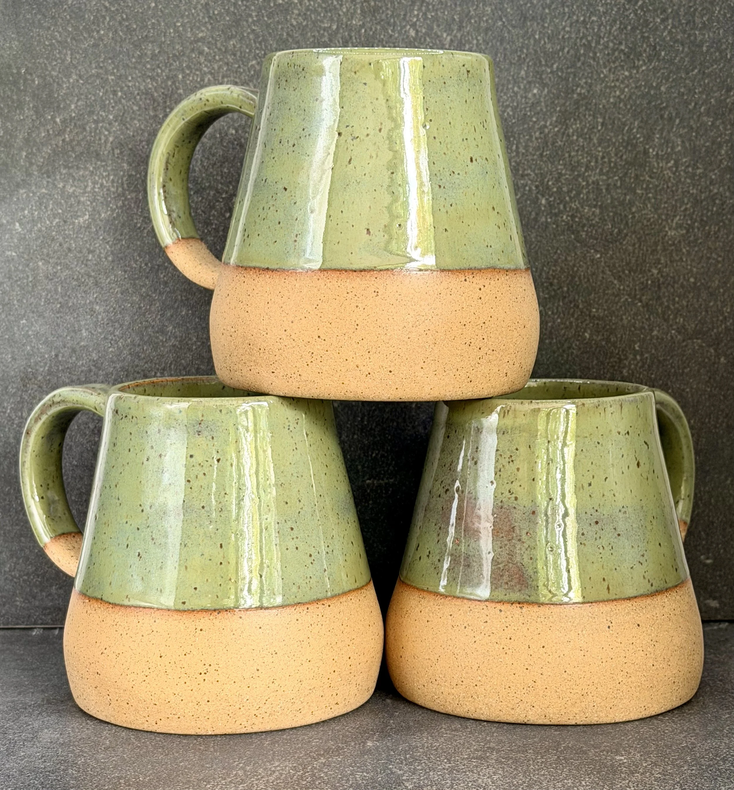 Hand built, wide-bottom ceramic mugs with a rustic, speckled clay bottom and a green, glossy glazed upper section, stacked on top of each other on a dark, textured surface.
