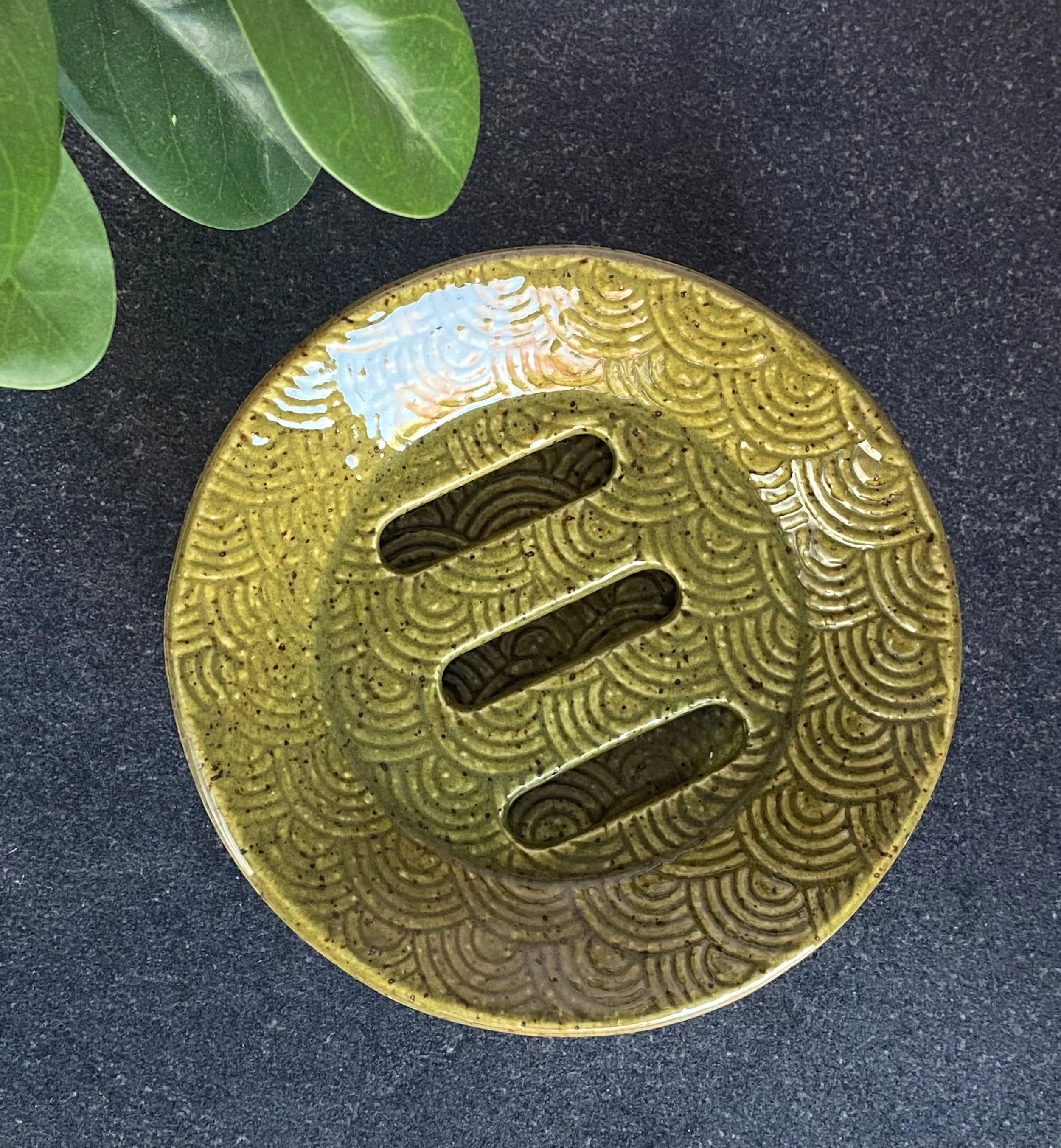 Close-up of a round, speckled olive green ceramic soap dish with intricate rainbow scale pattern on a dark countertop. Includes drainage tray.