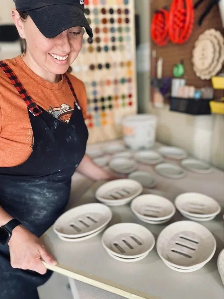 A woman in a black cap and orange t-shirt smiles while working with ceramic bowls on a table, some with slotted openings.