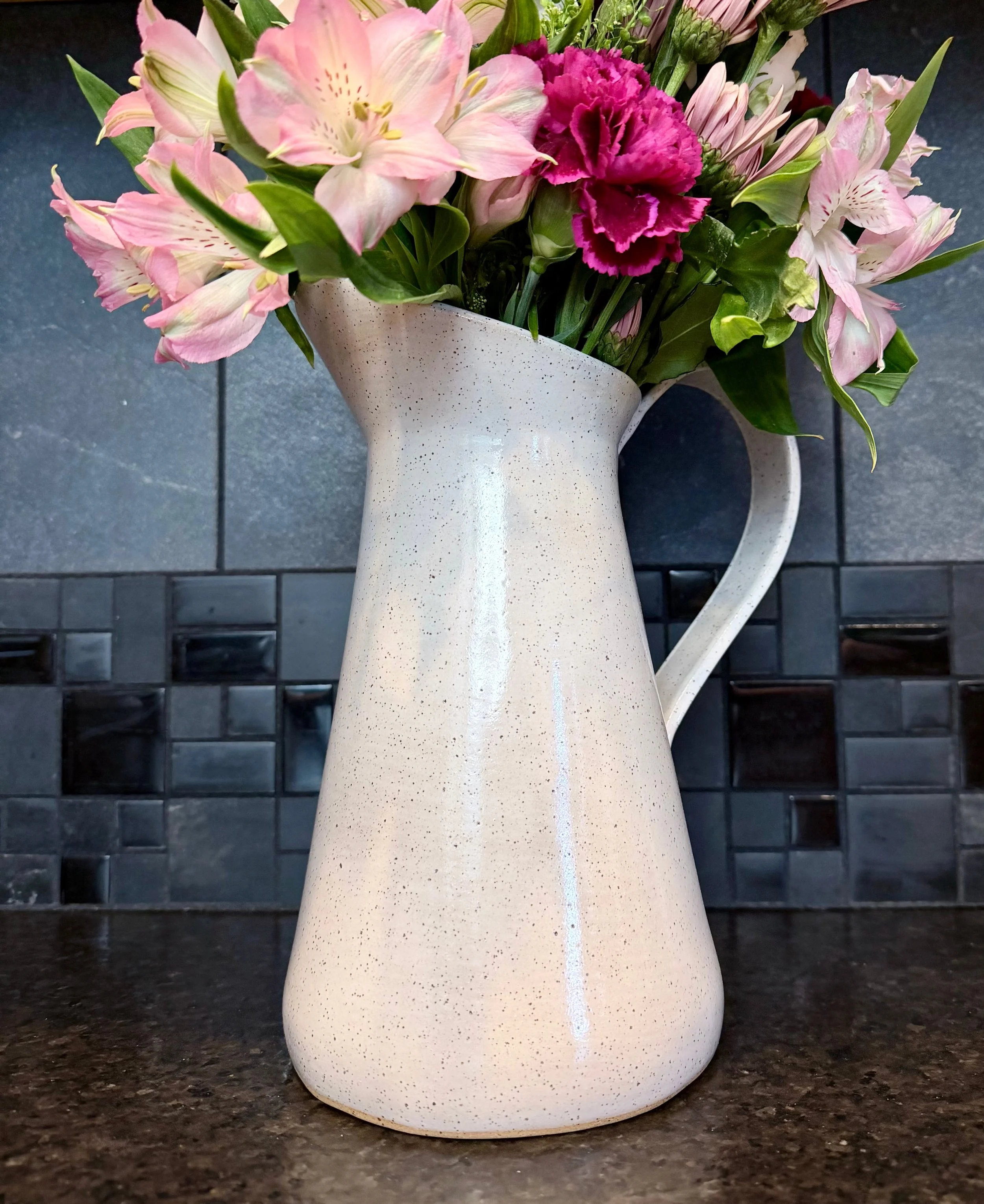 A ceramic hand-built vase and pitcher with a speckled off-white finish holding a bouquet of pink and purple flowers, set on a dark granite countertop with a dark tiled backsplash.