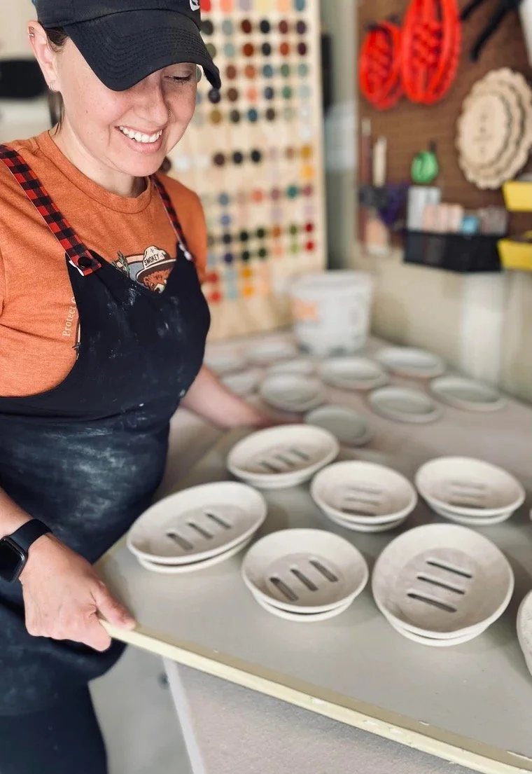 A woman in a black cap and apron holding a tray of unglazed ceramic bowls with knife marks inside, working in a pottery studio.