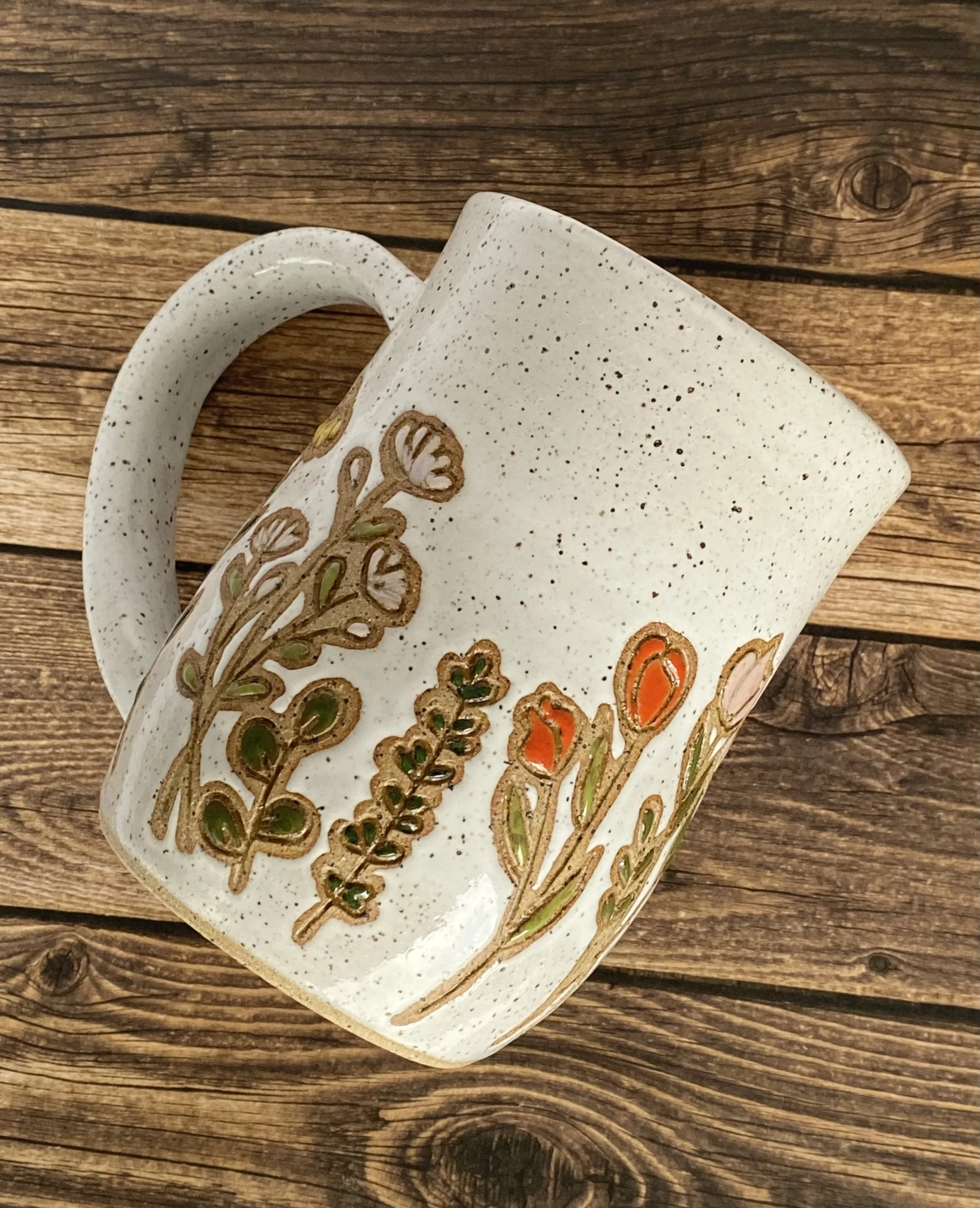 A ceramic mug with a white-colored speckled glaze and a decorative hand-carved floral design, resting on a wooden surface.
