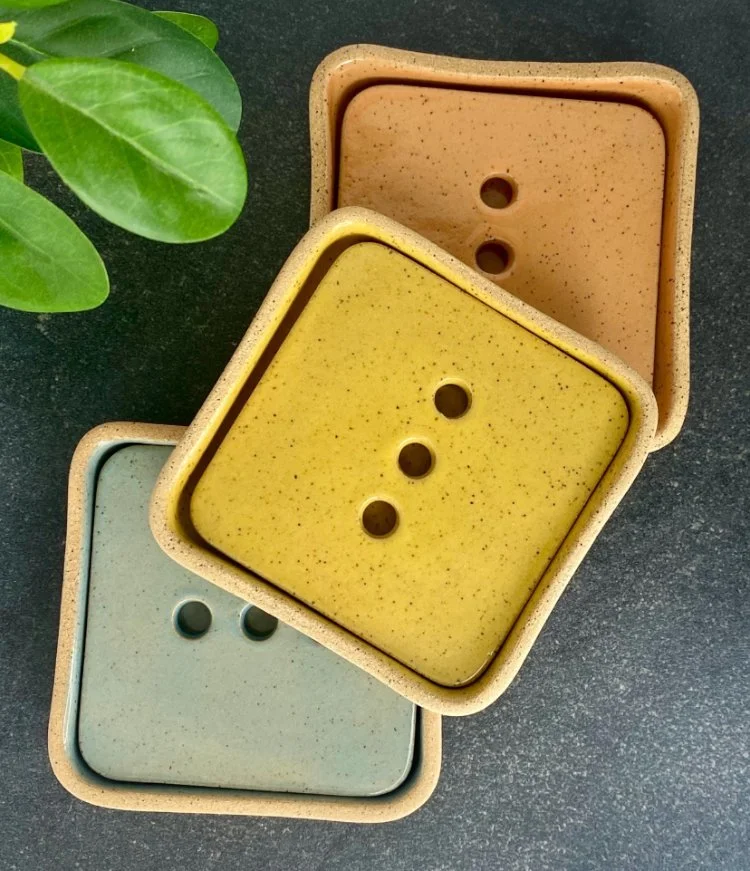 Three square ceramic soap dishes with round openings, stacked and placed on a dark surface with green leaves nearby.