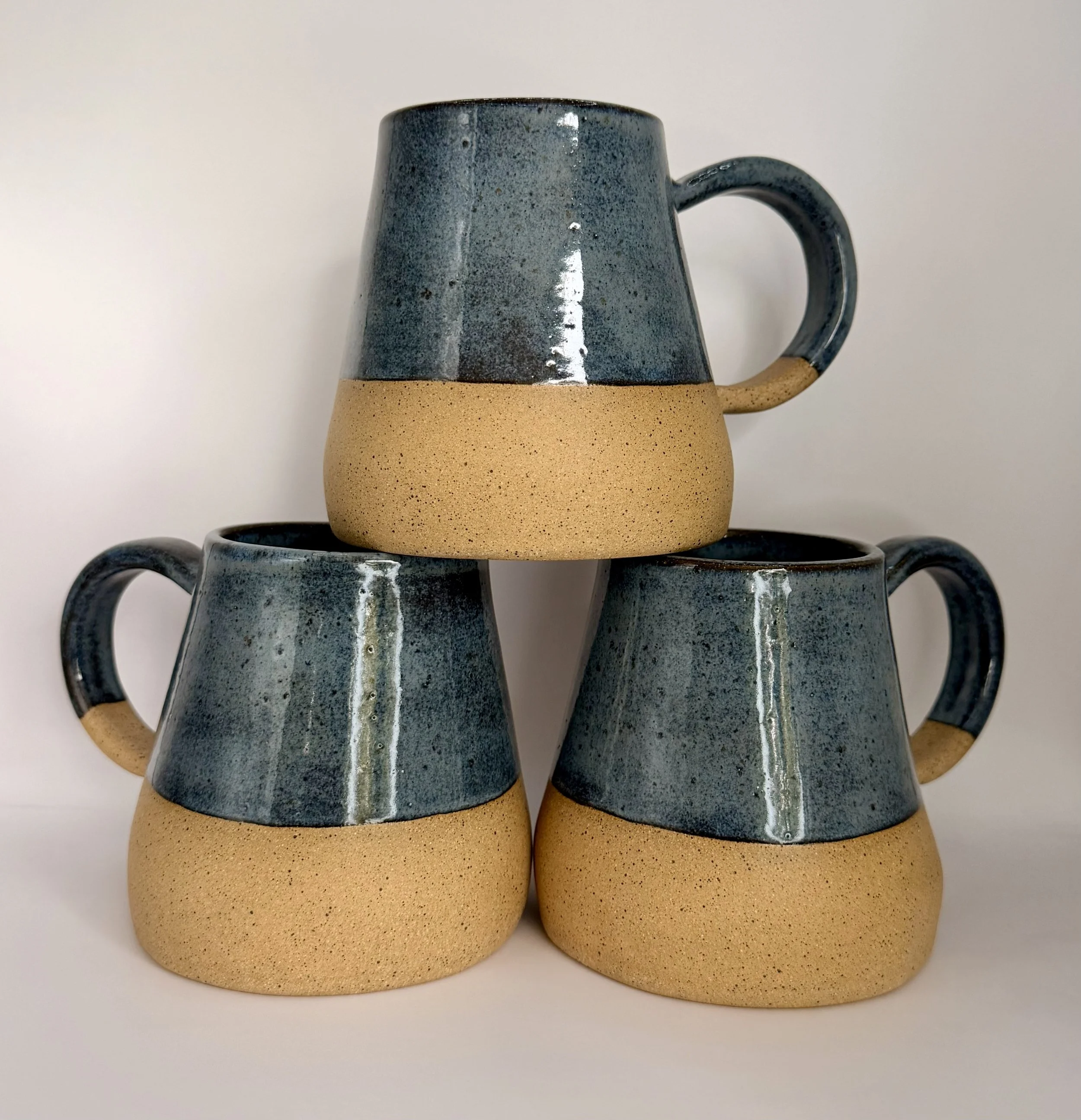 A stack of three blue ceramic hand-built wide-bottom mugs.