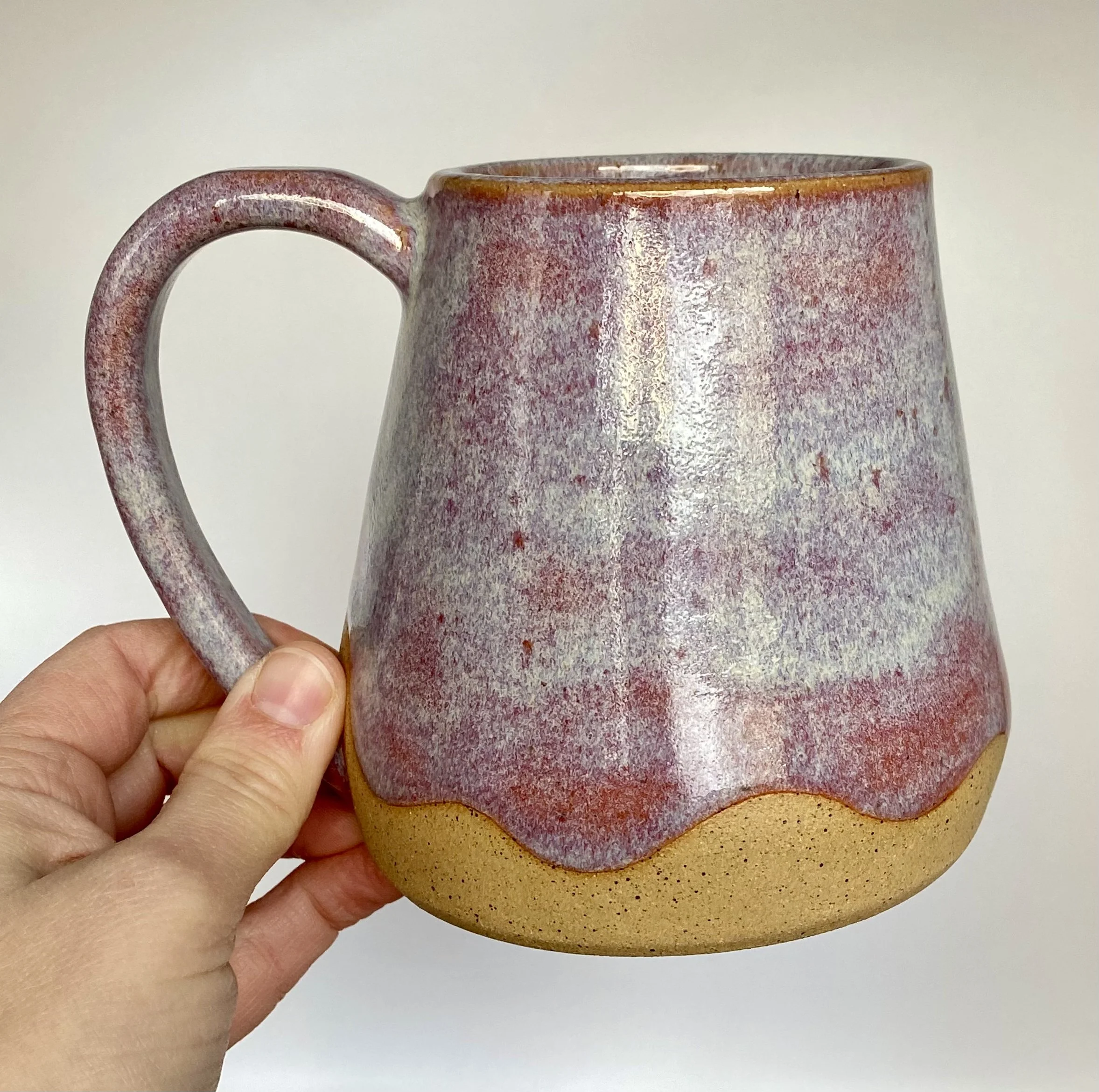 Close-up of a hand-built wide-bottom ceramic mug with a purple, blue, and red glaze pattern, being held in a person's hand.