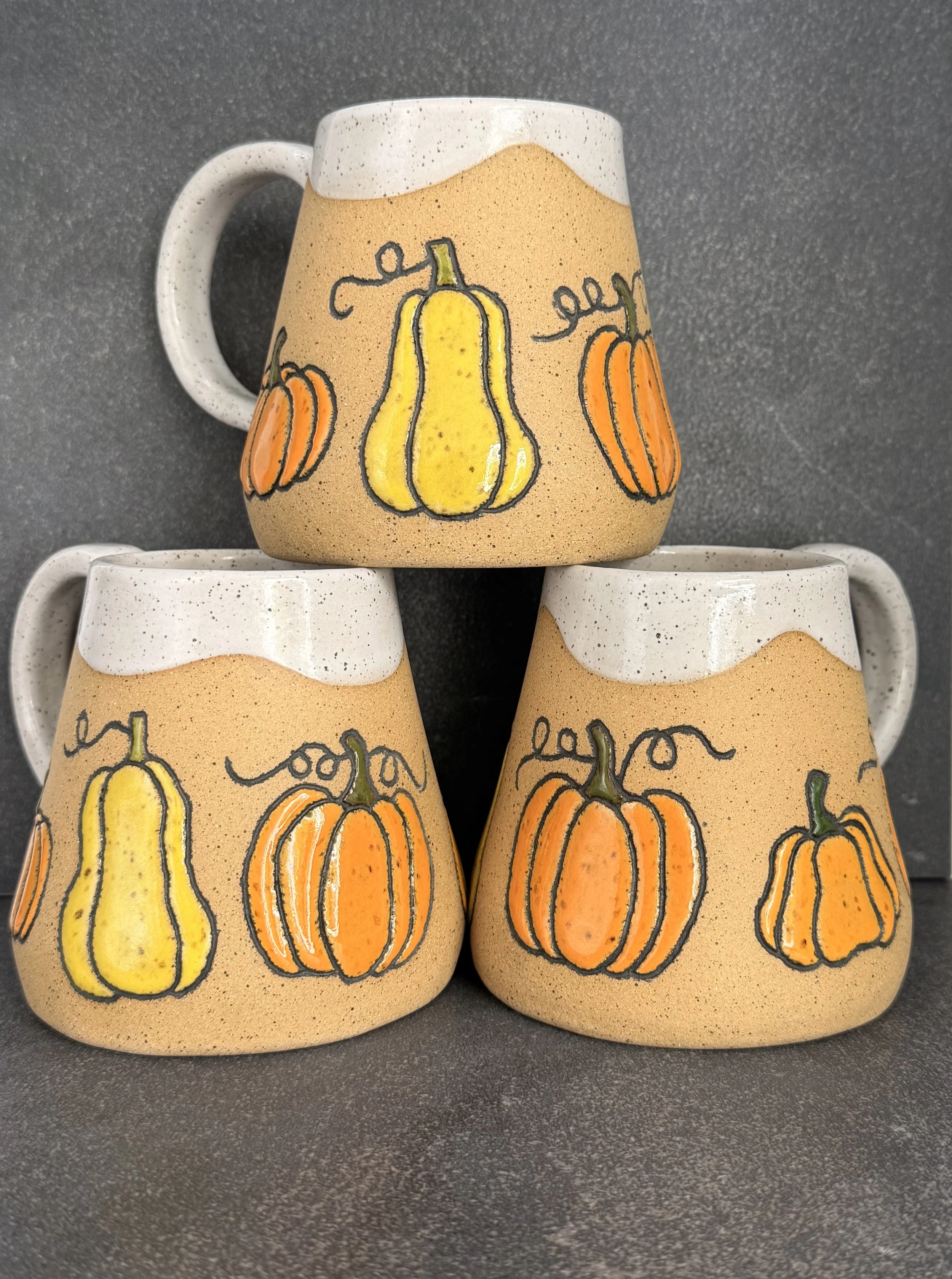 Three hand built ceramic mugs with a pumpkin-themed decoration, featuring a large yellow and an orange pumpkin with green stems, placed on a dark gray surface.