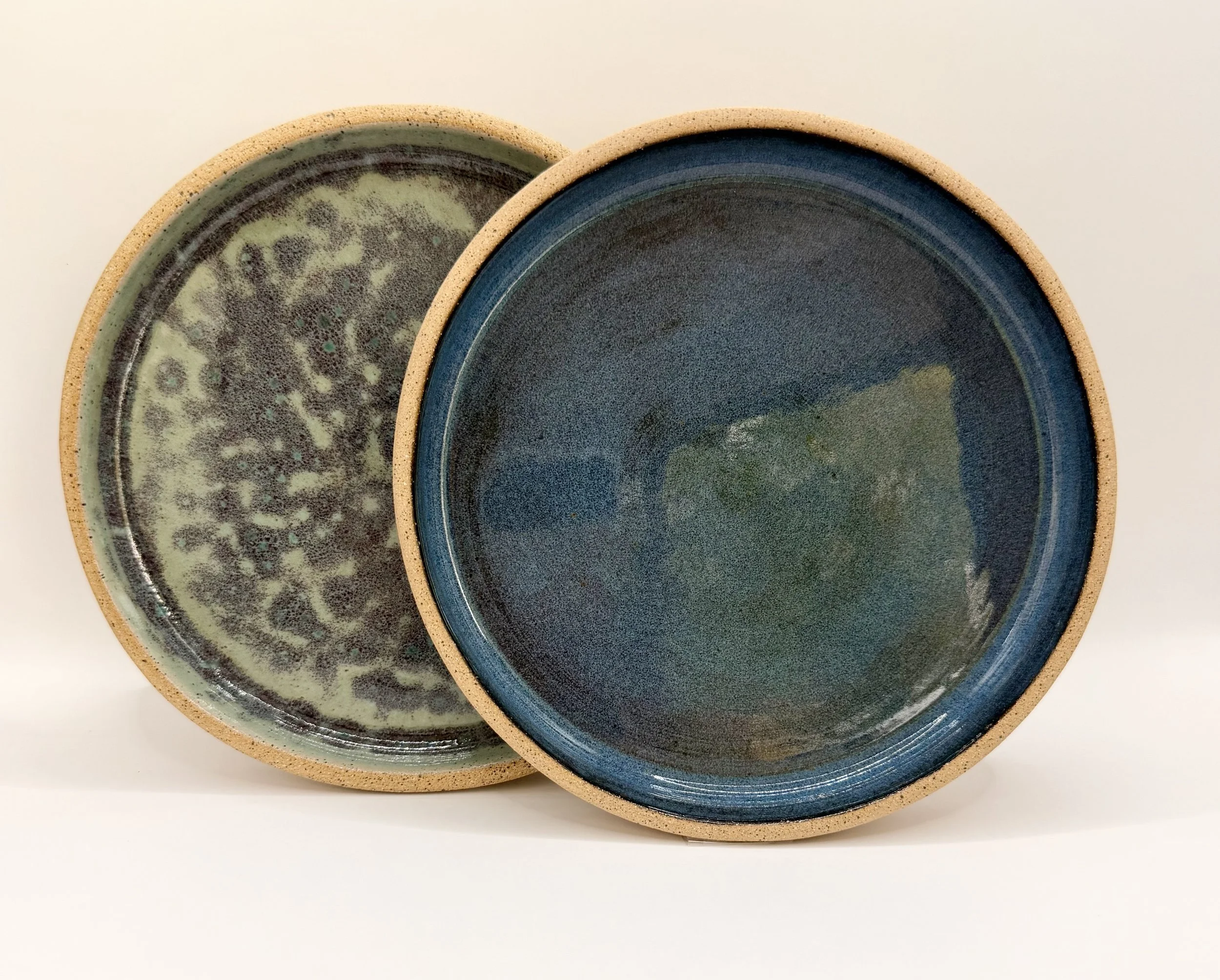 Two overlapping 8" ceramic plates with blue and green glazing, showing a speckled pattern and a raw speckled clay rim, placed on a white surface.