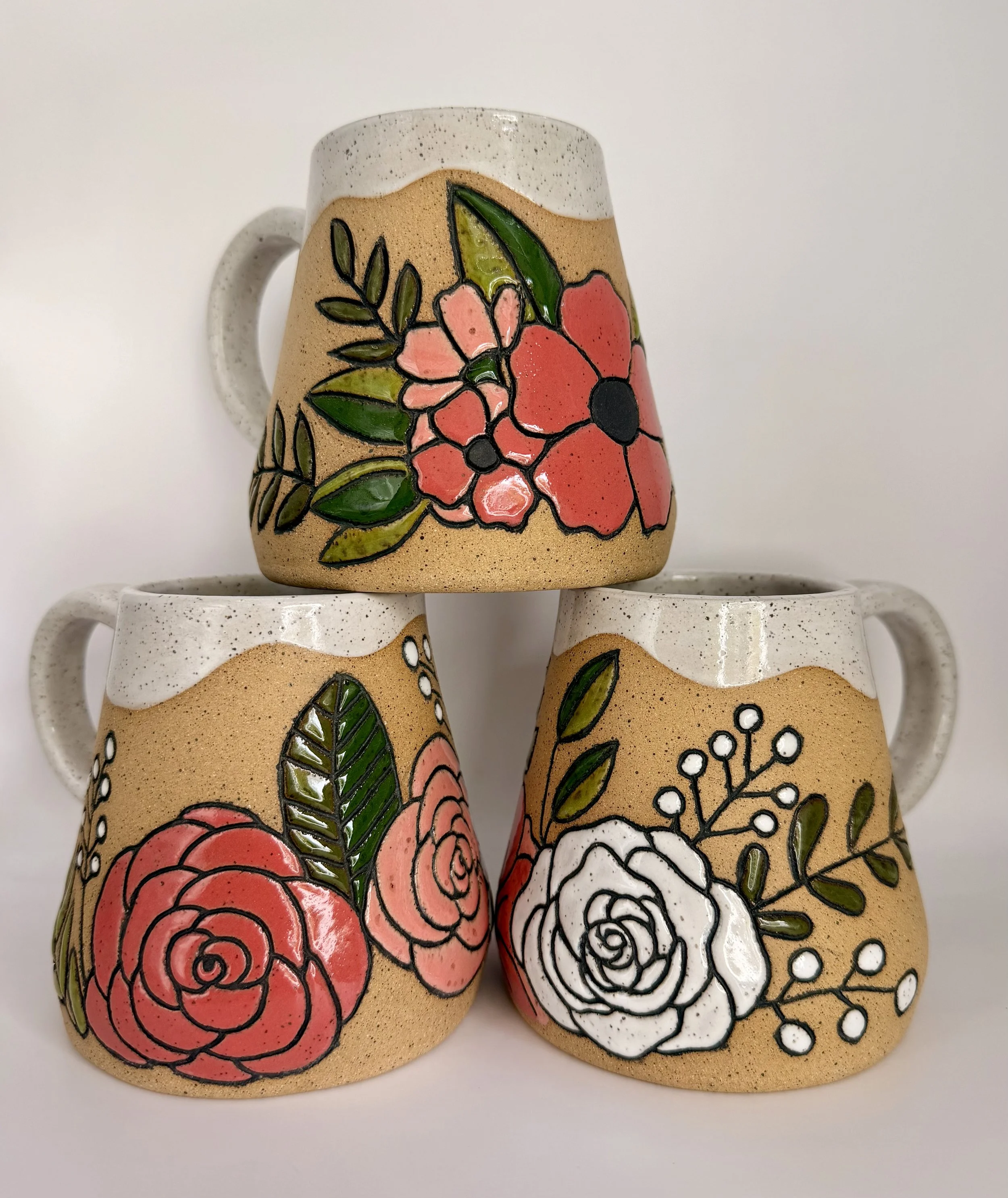 Three hand-built wide-bottom ceramic mugs with painted floral designs, featuring red and white roses with green leaves, positioned against a plain white background.