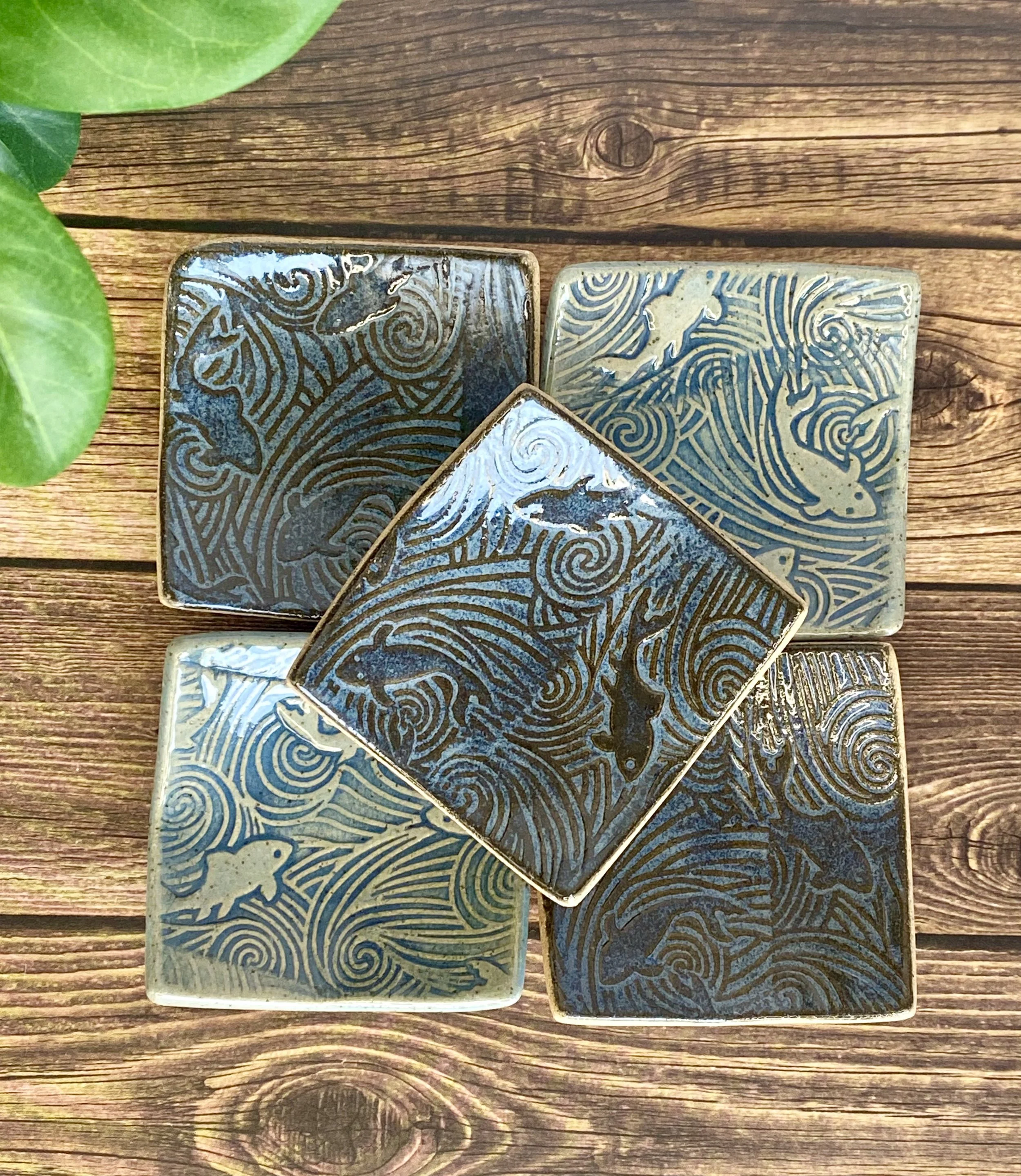 A close-up of five decorative jewelry and trinket dishes with intricate figh patterns, arranged on a wooden surface with green leaves partially visible in the top left corner.