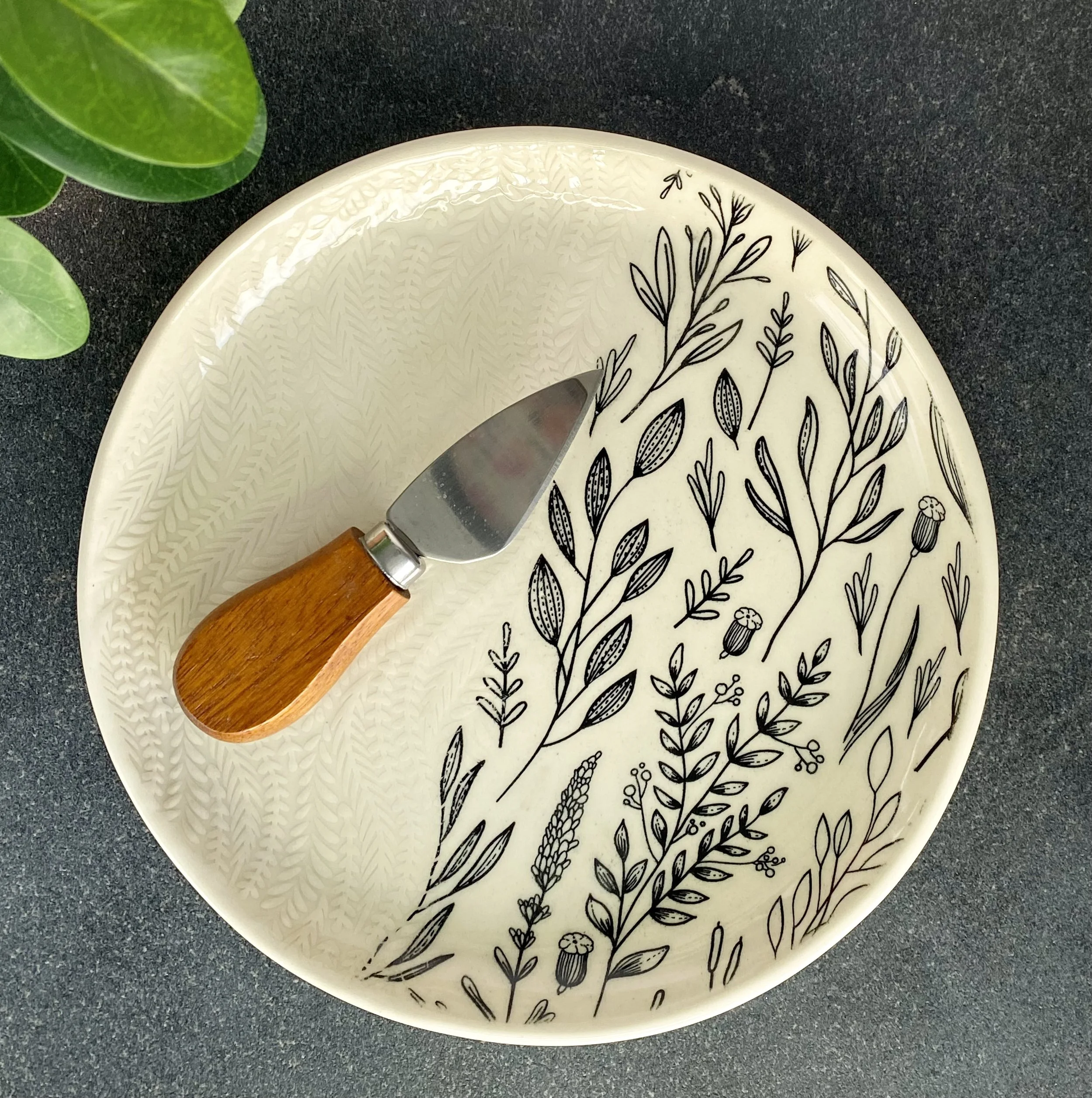 An off-white ceramic plate with black leaf and plant patterns, a knife with a wooden handle resting on it, and a green plant with broad leaves on a dark textured surface.
