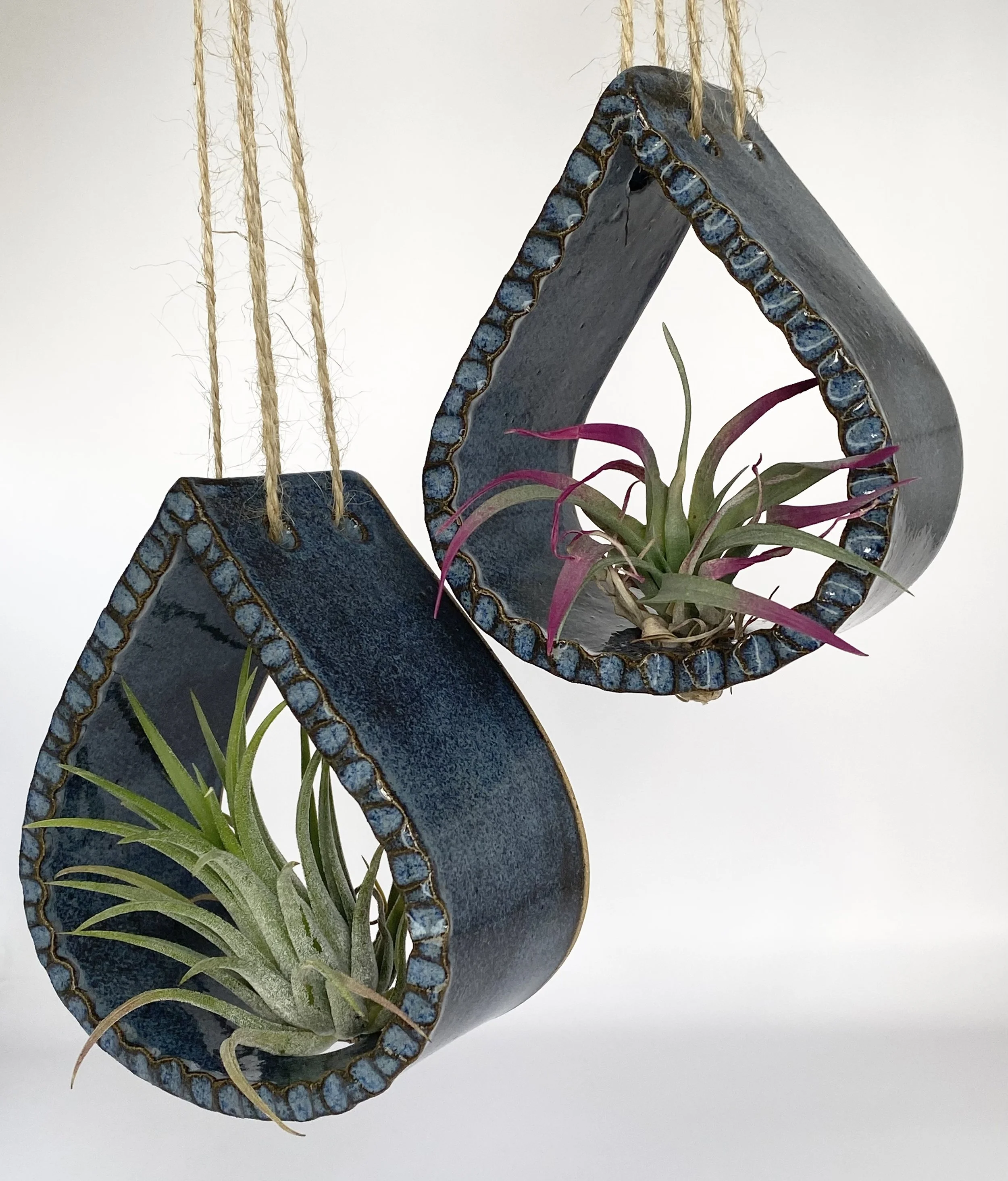 Decorative blue ceramic airplant holder shaped like a teardrop, with a hole in the center filled with a small green and purple plant, hanging from jute ropes against a plain white background.