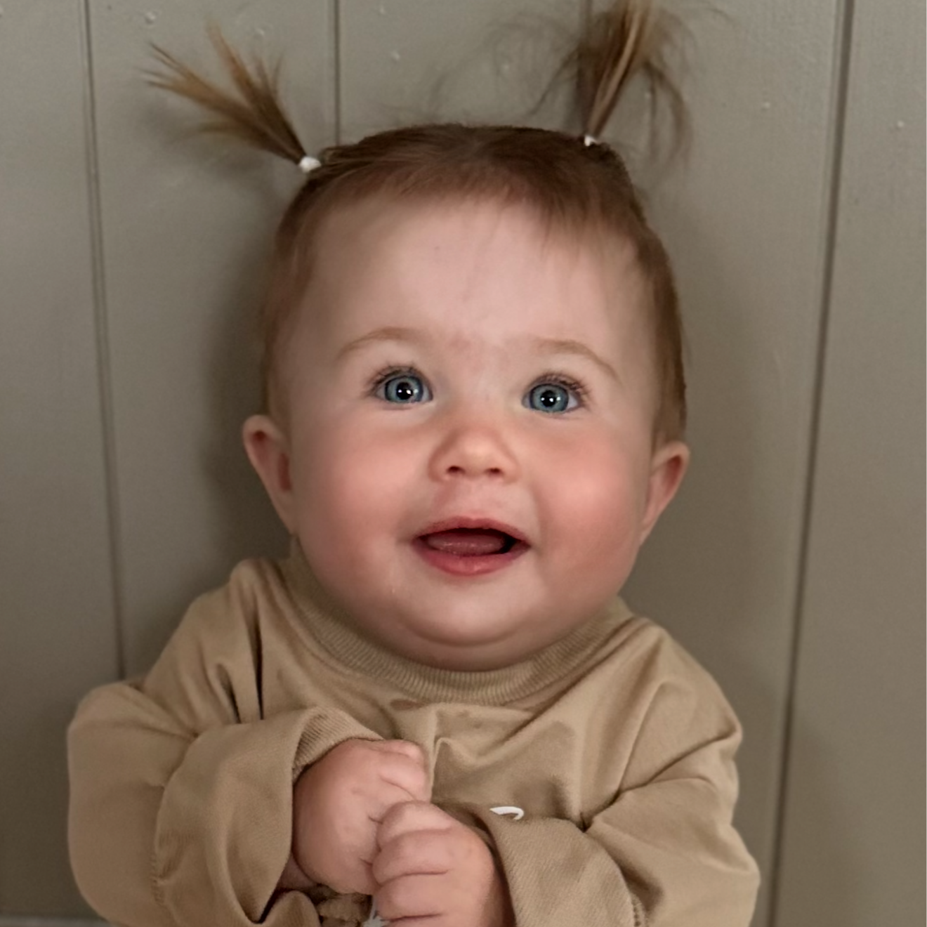 A young child with blue eyes and red hair styled in two pigtails, smiling and wearing a beige long-sleeve shirt, standing against a gray paneled wall.