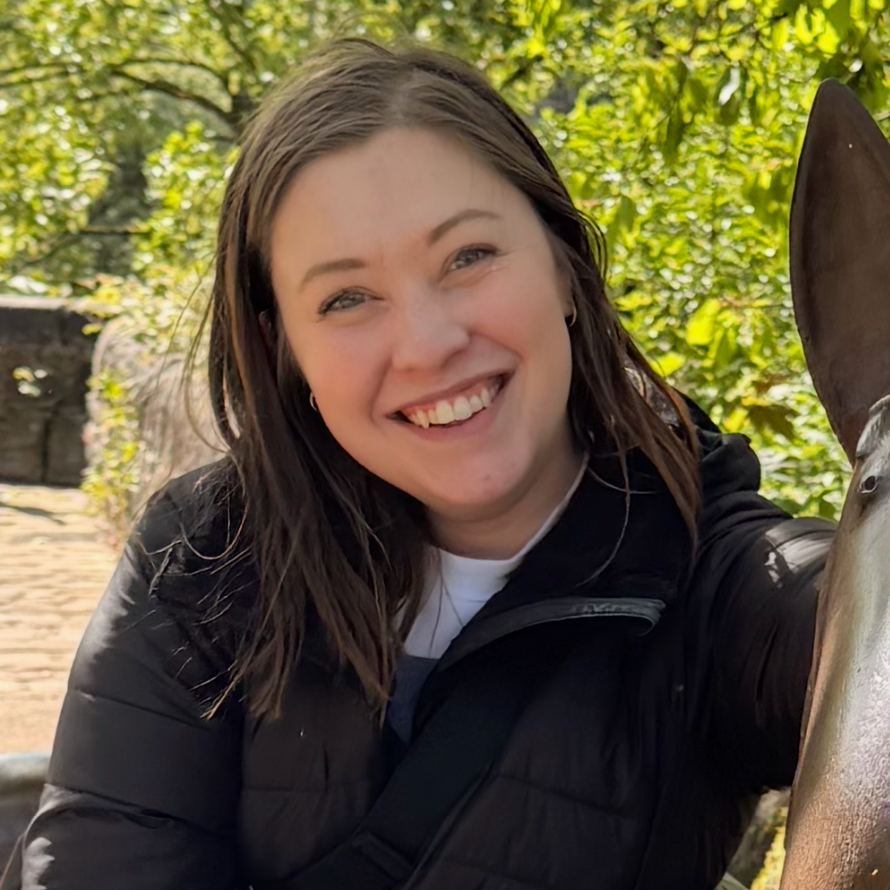 A woman with brown hair smiling outdoors next to a horse.