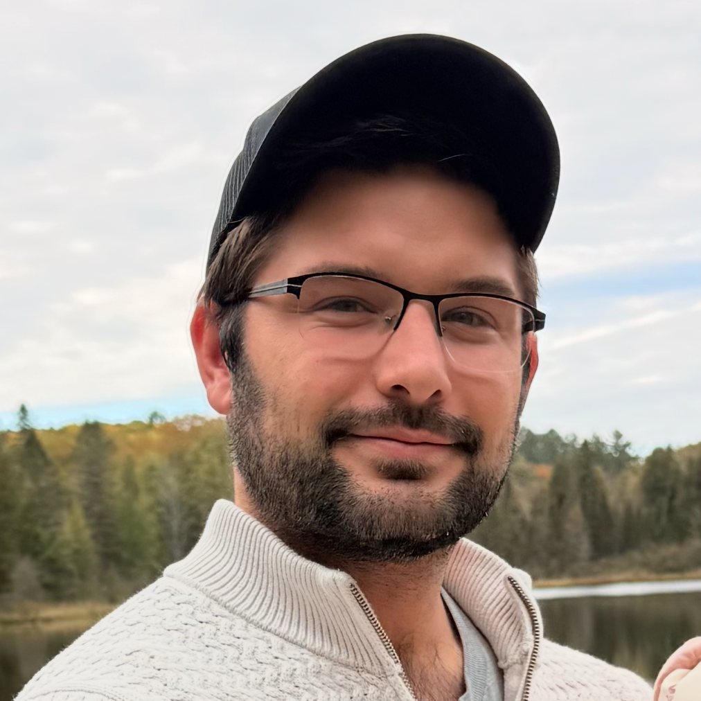 A man with glasses, a baseball cap, and a beard posing outdoors with a lake and trees in the background.