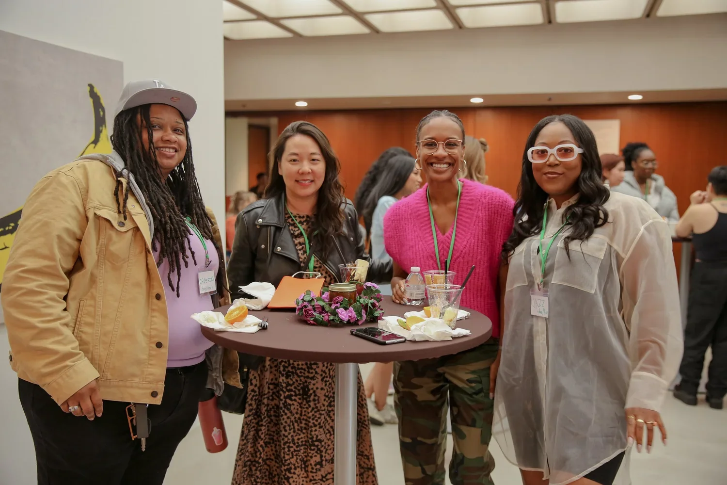 Four women standing around a high table at a social event. The table has drinks, snacks, a small flower arrangement, and a phone. The women are smiling and wearing casual attire with name tags. The background shows a busy event space.