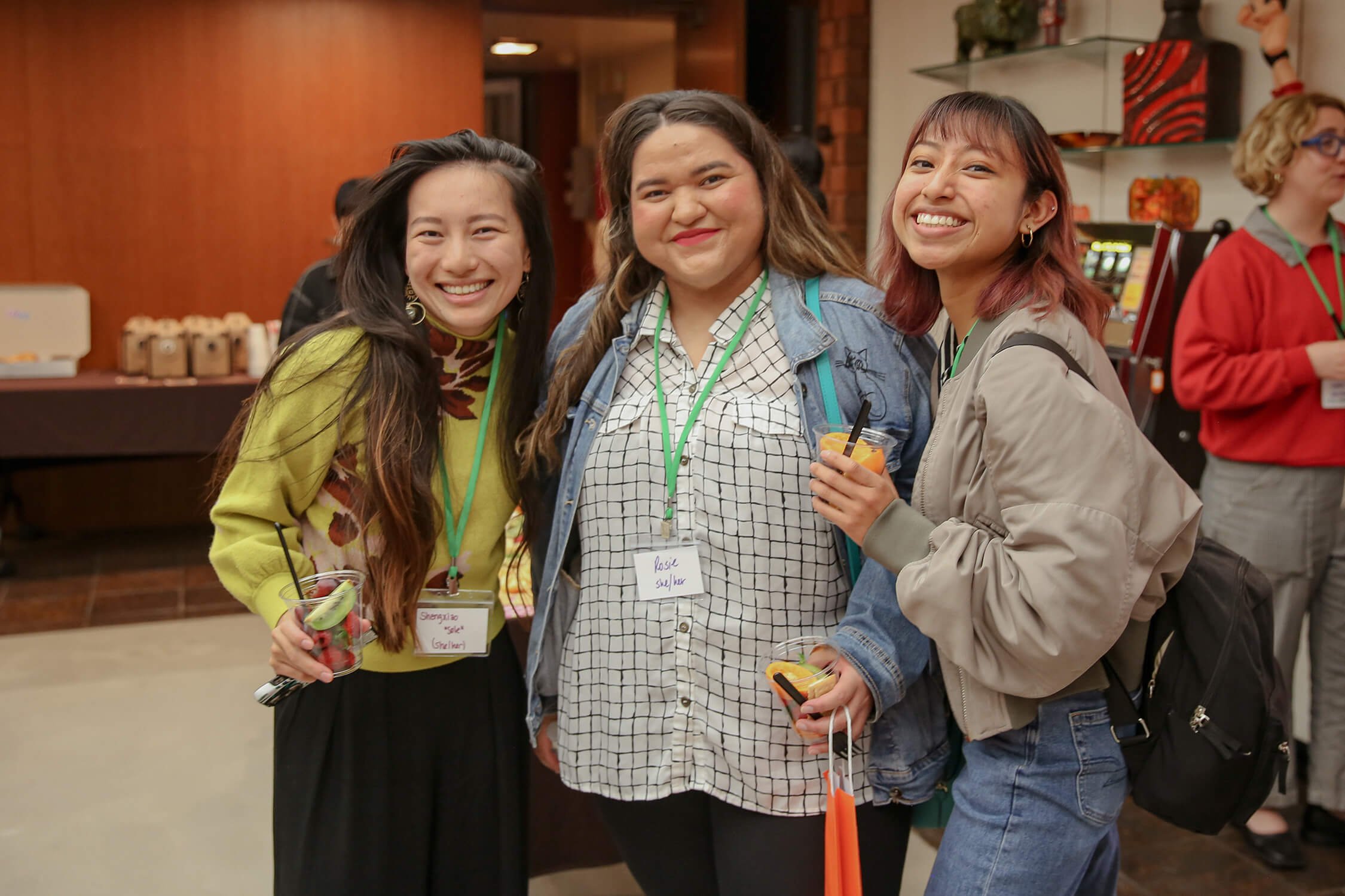 Three women smiling and posing together at an indoor event, each holding a drink in their hands. They are wearing name tags with one reading "Rosie" and others partially visible. The background shows a wooden wall and a counter with cups and beverages.