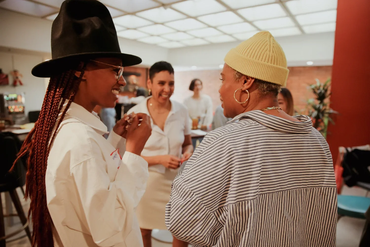 Six people socializing indoors, two women in the foreground shaking hands and smiling, others in the background holding drinks and talking.