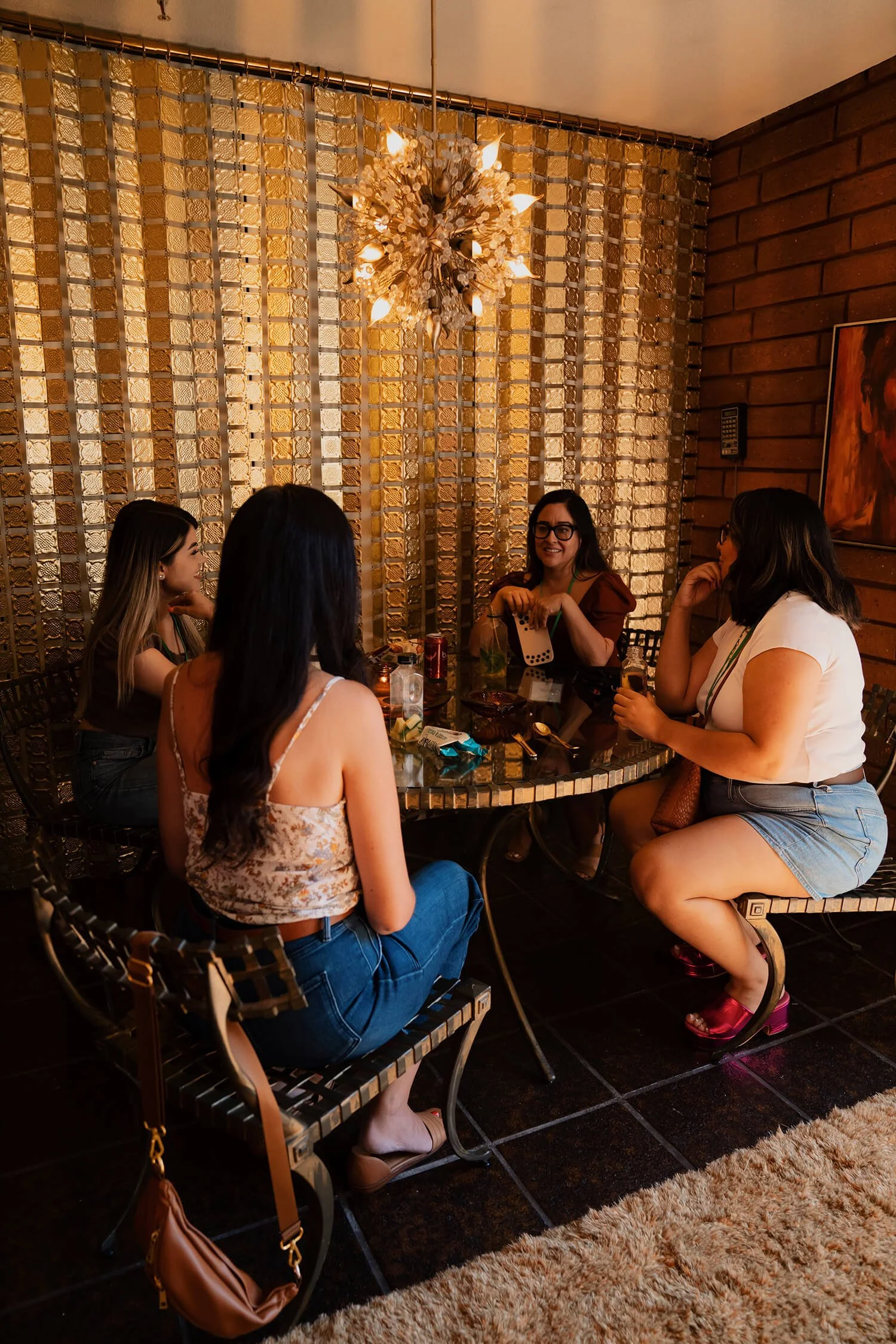 A group of six women sitting around a round table in a dimly lit restaurant or bar, with a decorative wall, chandelier, and warm lighting.