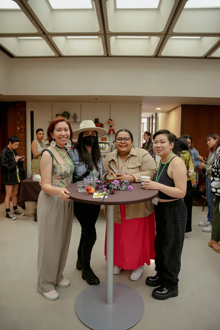 Four women standing around a high table at a social gathering, smiling at the camera. They are indoors, with other people in the background. The table has flowers, drinks, and snacks on it.