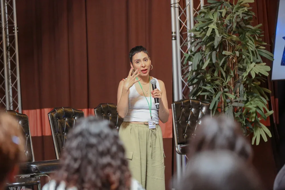 Woman with short dark hair, wearing a white tank top and beige pants, speaking into a microphone at a conference or seminar, with an audience and potted plant in the background.