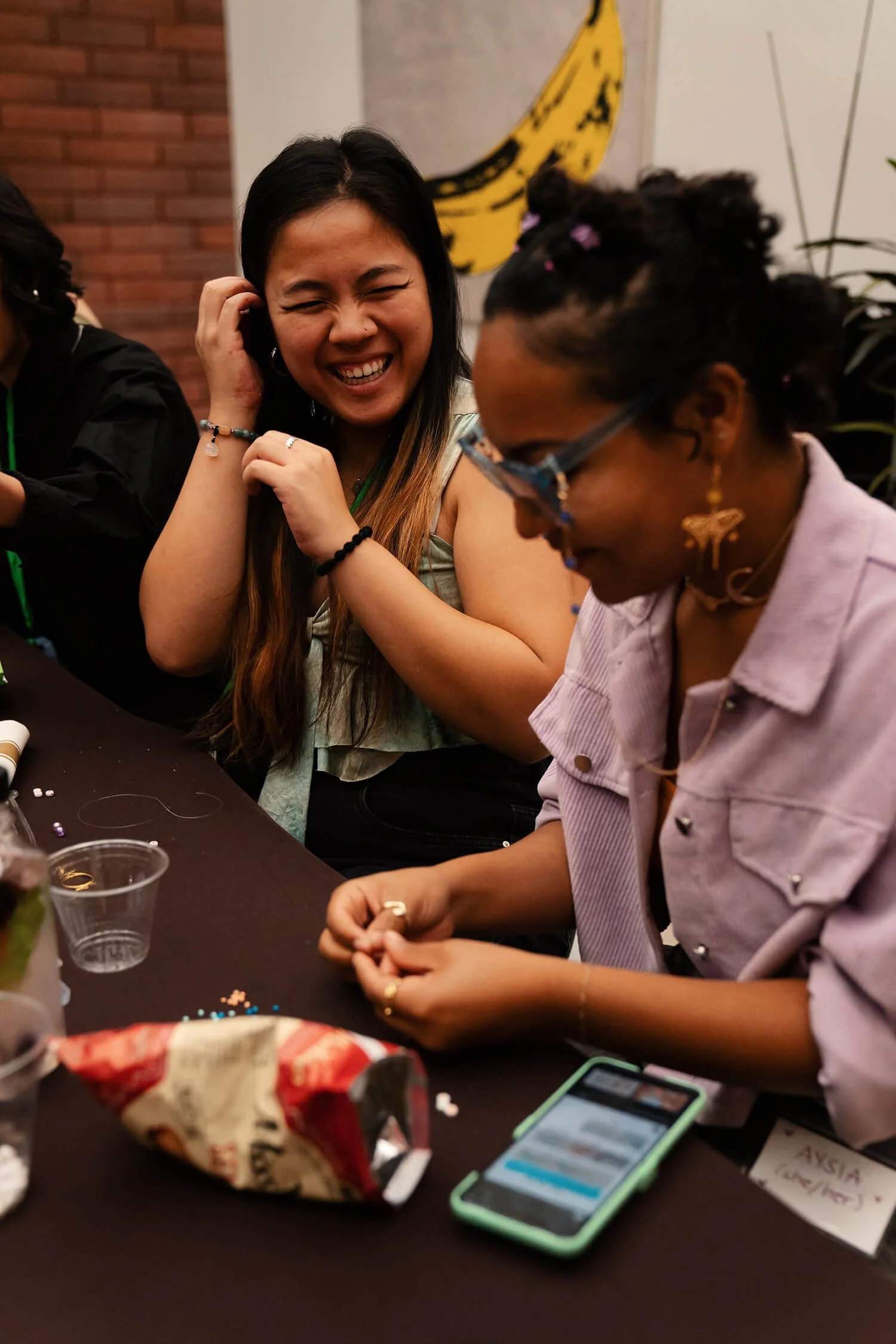 Two women sitting at a table, one with curly hair and glasses, the other with long dark hair, both smiling and looking down. The table has various items, including a smartphone, jewelry, and snacks. A banana drawing is on the wall in the background.