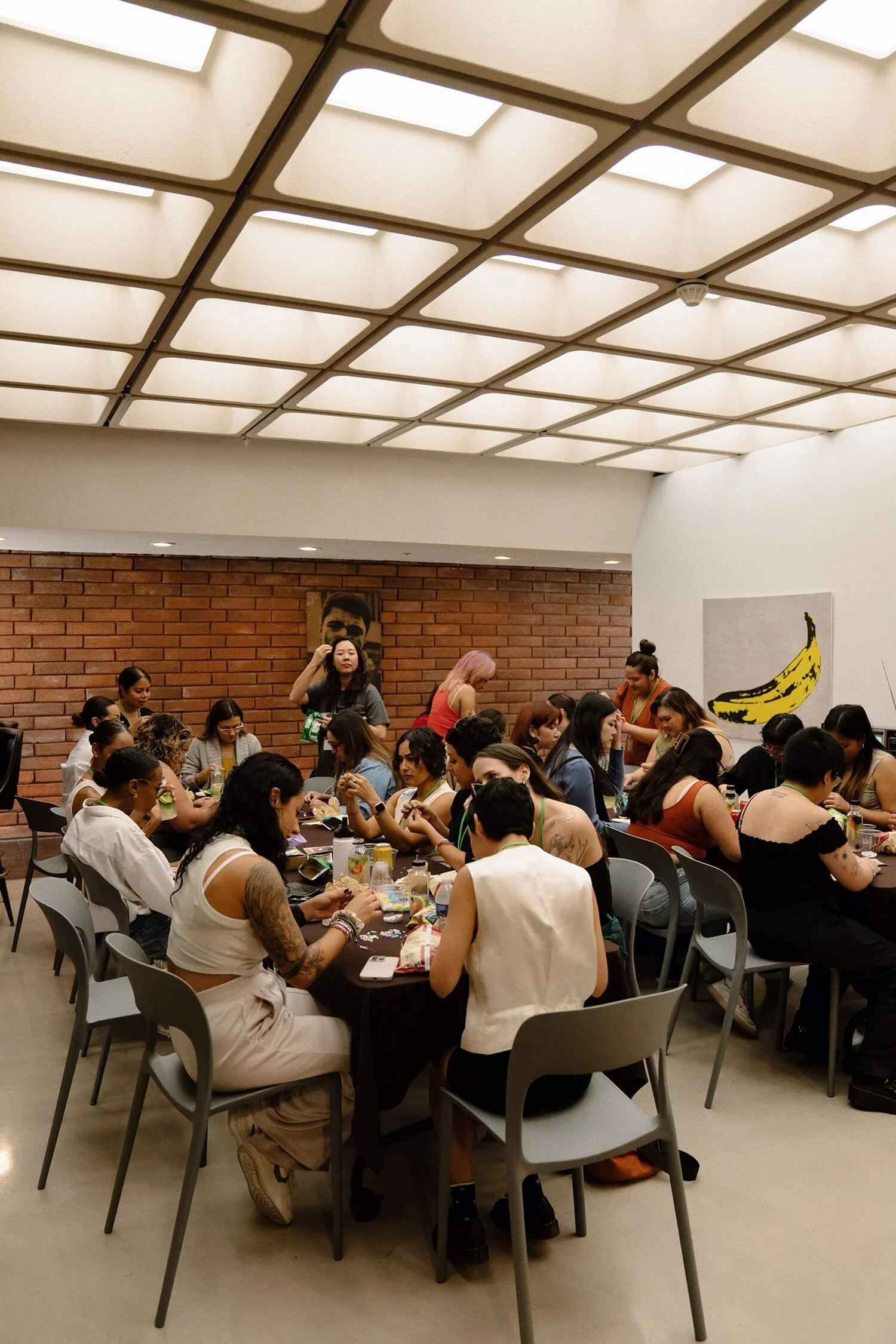 A group of women gathered around a large table in a room with a brick wall, some are working on arts and crafts while others are talking, and a portrait hangs on the wall.
