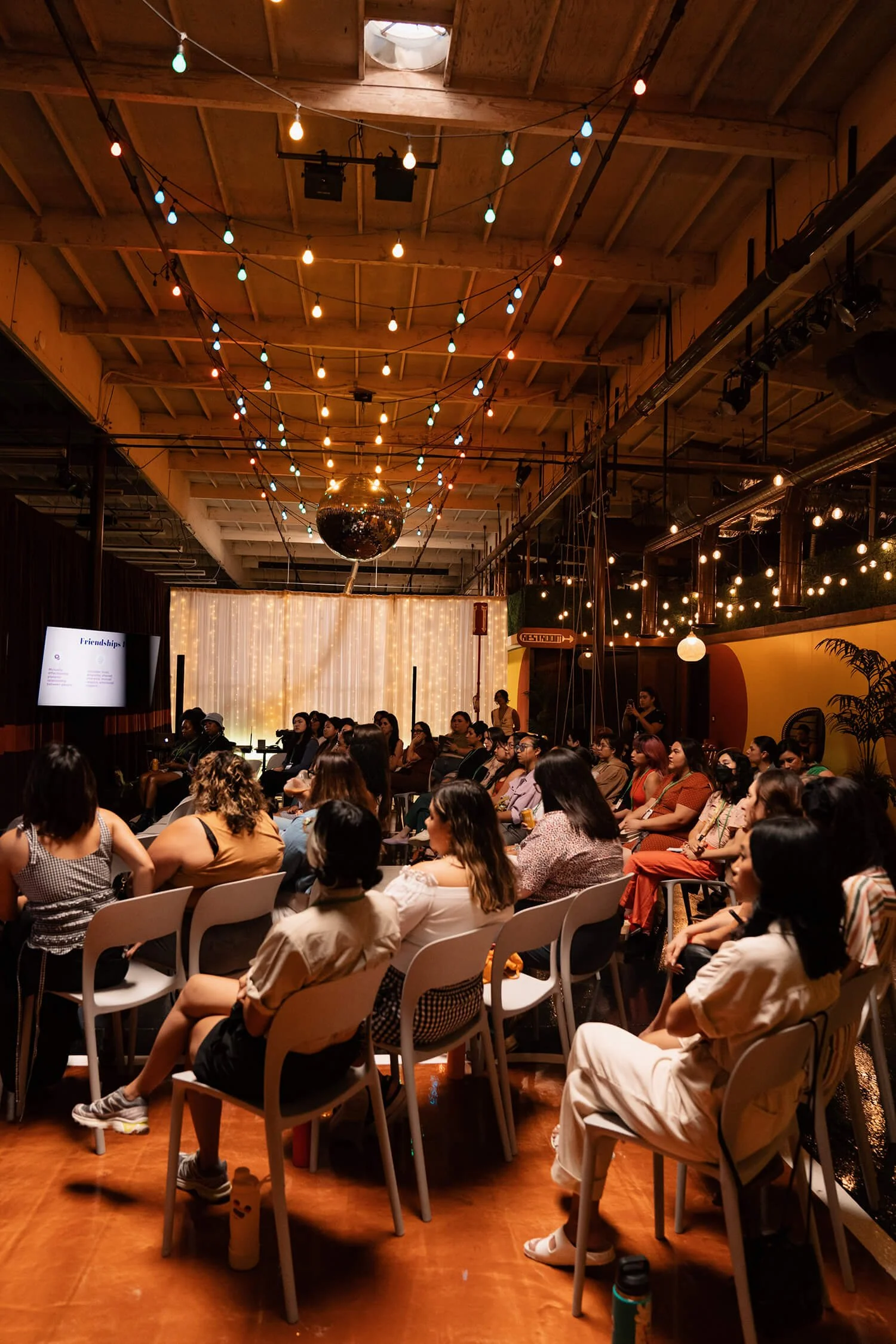 A large group of people seated in rows of chairs inside a decorated event space with hanging lights and a disco ball hanging from the ceiling, attending a presentation or performance.