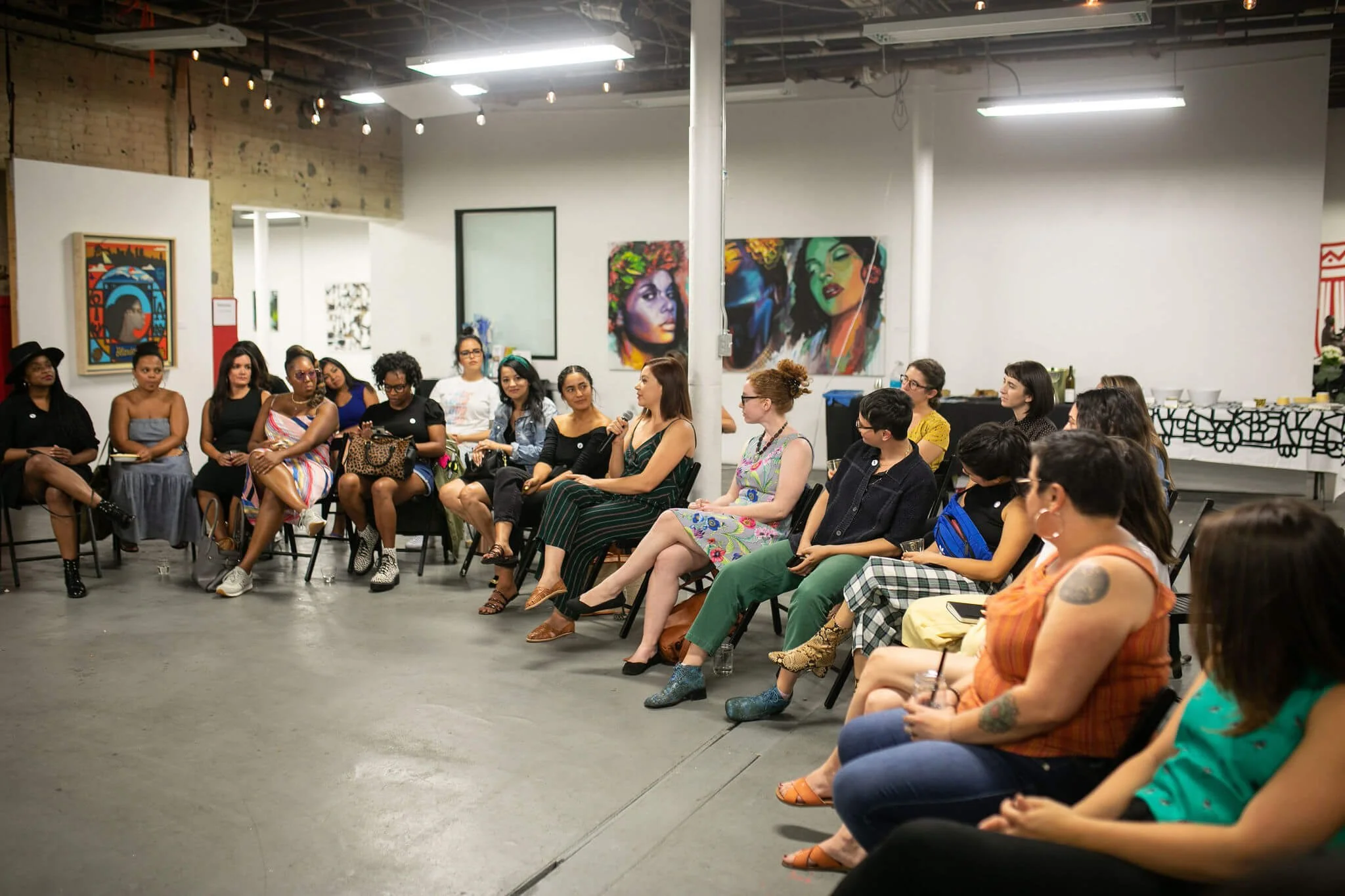 A diverse group of women participating in a discussion or panel in an art gallery or studio, with colorful paintings on the walls.