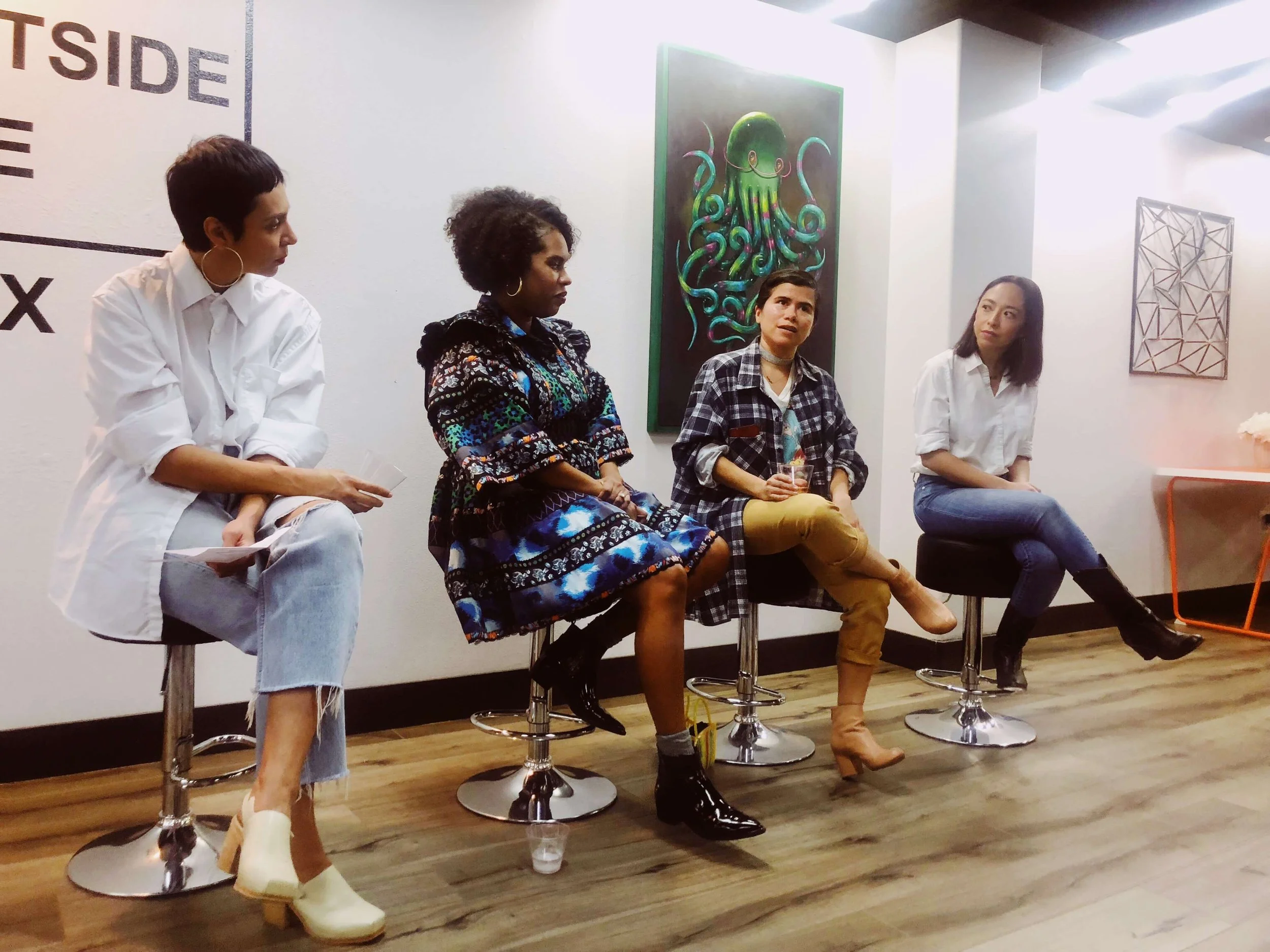 A group of four diverse women sitting on bar stools in a modern art gallery, engaging in a discussion. There are paintings on the wall behind them, and one woman is holding a drink. The room has wooden floors and white walls.