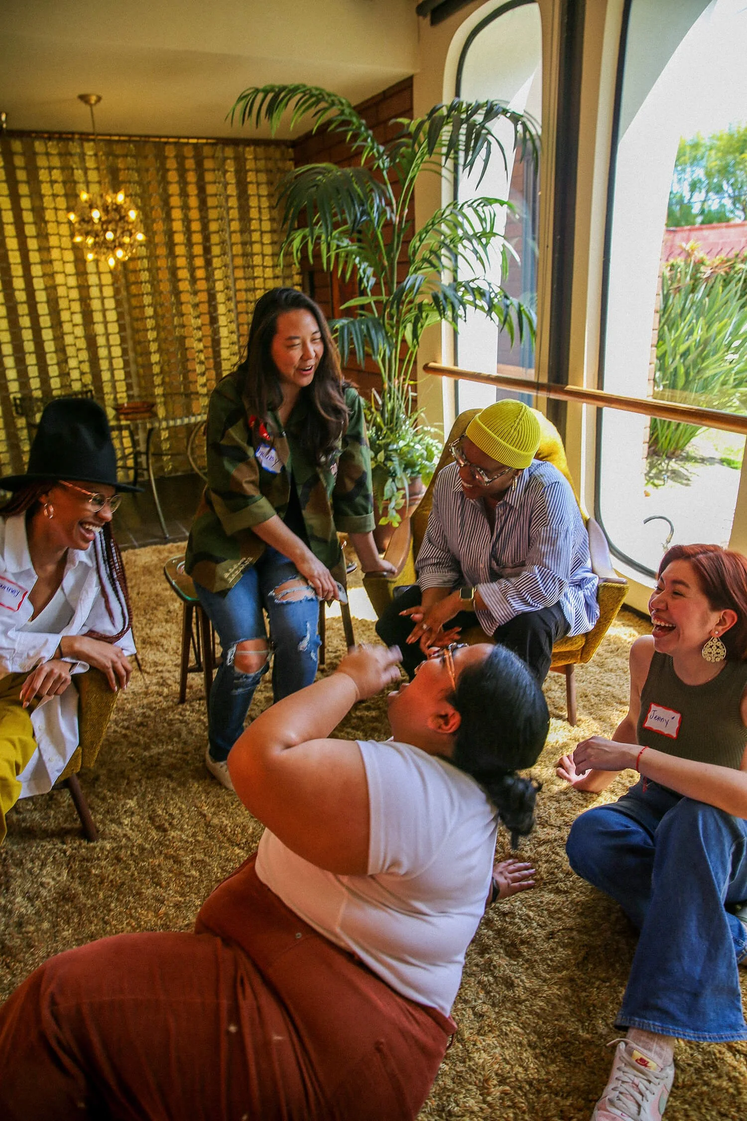A group of diverse people laughing and playing together indoors, some seated and some standing, near a large window with plants outside.