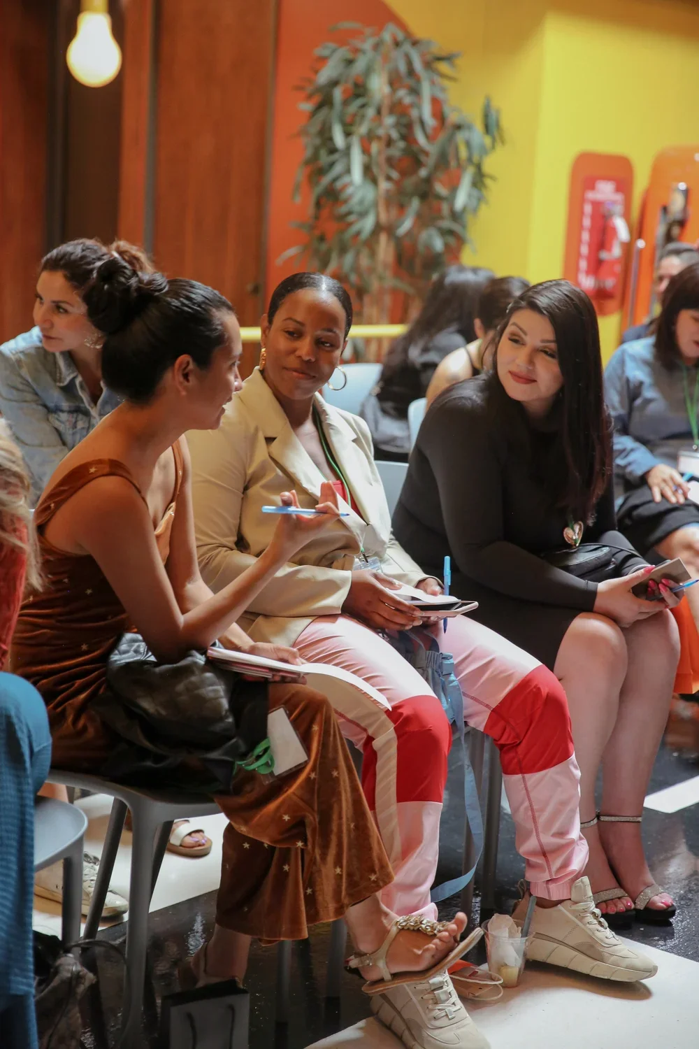 A group of women sitting in a colorful, casual indoor setting, engaged in conversation. Some are holding notebooks and pens, suggesting a seminar or workshop environment.