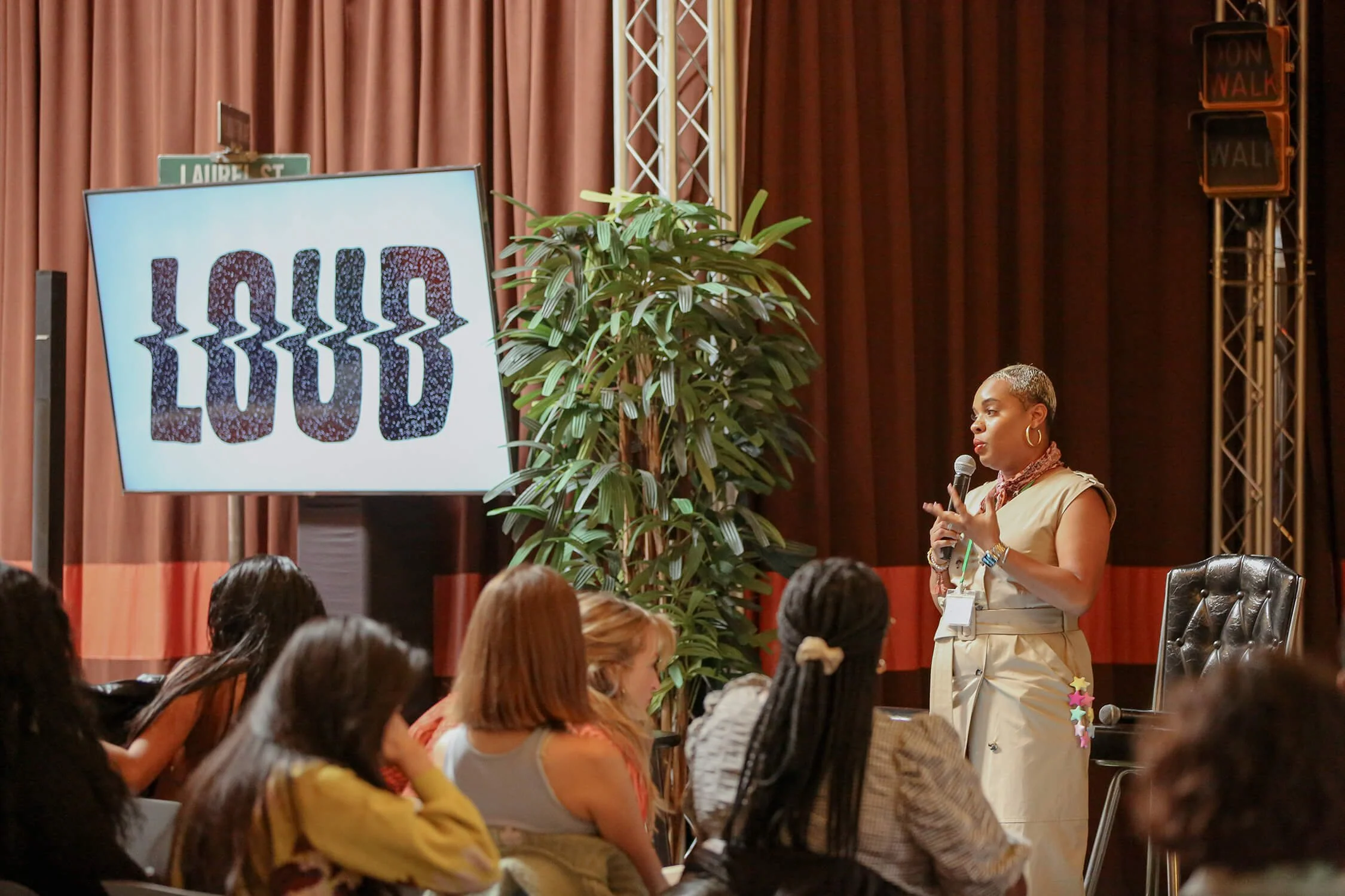 A woman standing and speaking into a microphone during a presentation to an audience, with a large screen displaying the word 'LOUD' and a city street sign in the background.