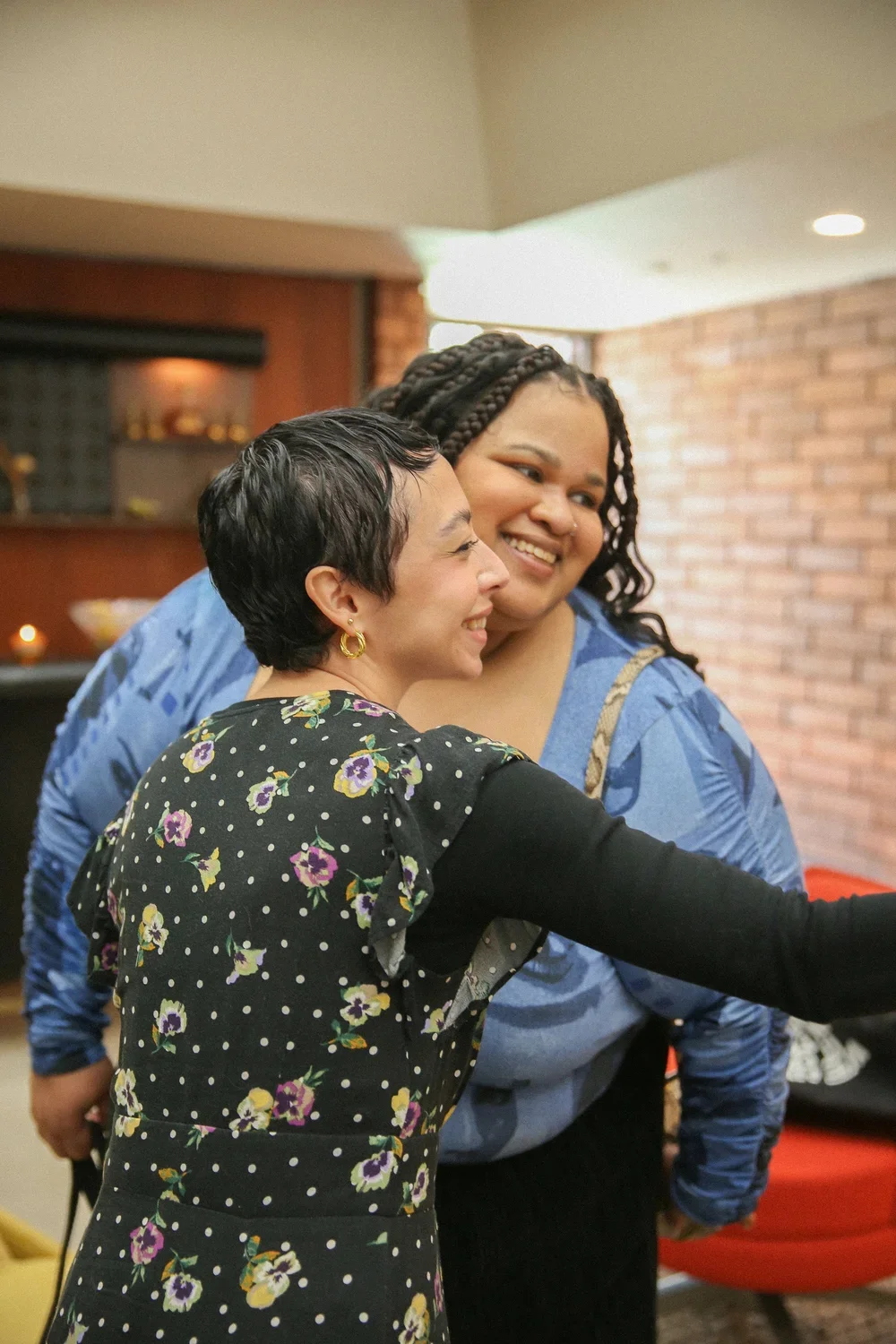 Two women smiling and hugging in a cozy indoor setting with brick walls and warm lighting.