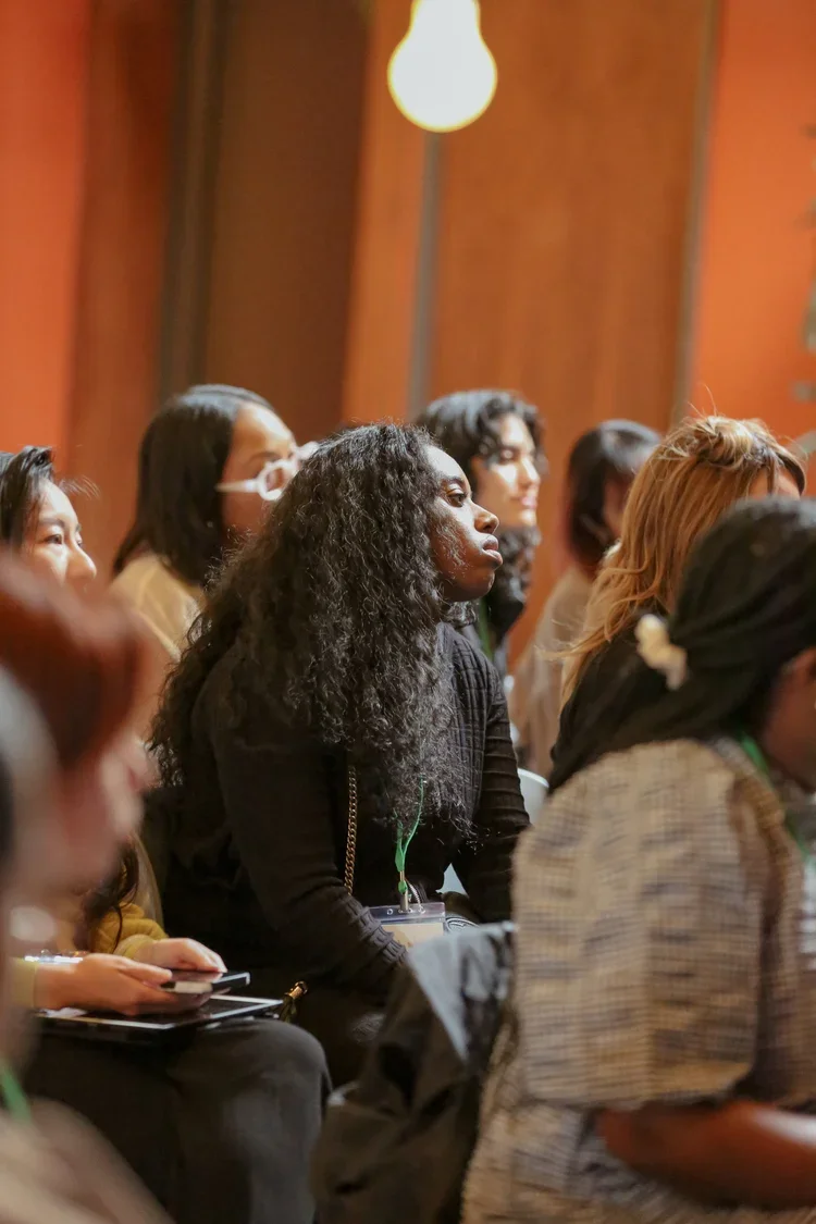 Group of diverse women attending a conference or seminar, seated and listening attentively, with a wooden wall and a hanging light in the background.