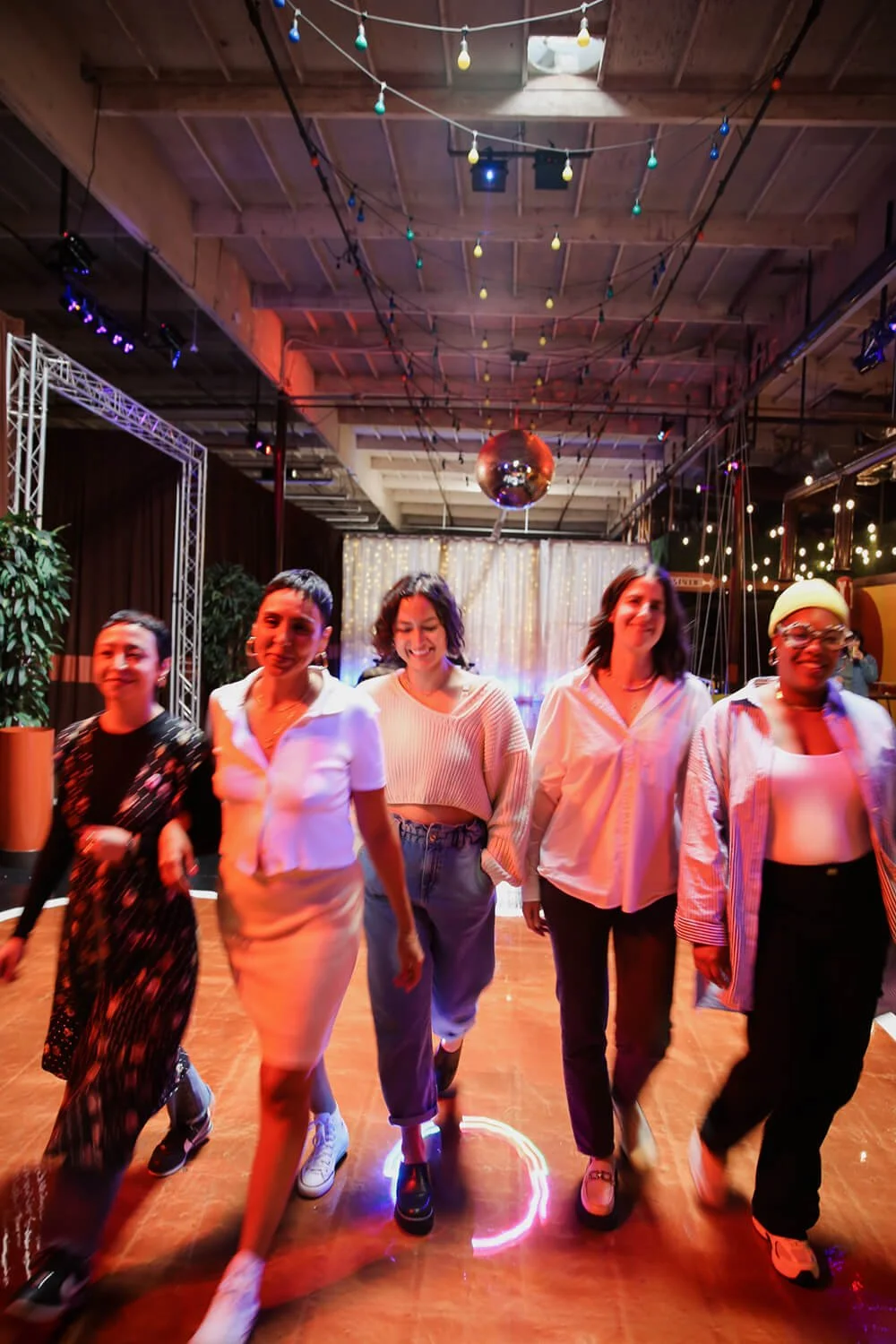 Five women walking together on a dance floor at a party or social event with colorful string lights, a disco ball, and decorative curtains in the background.