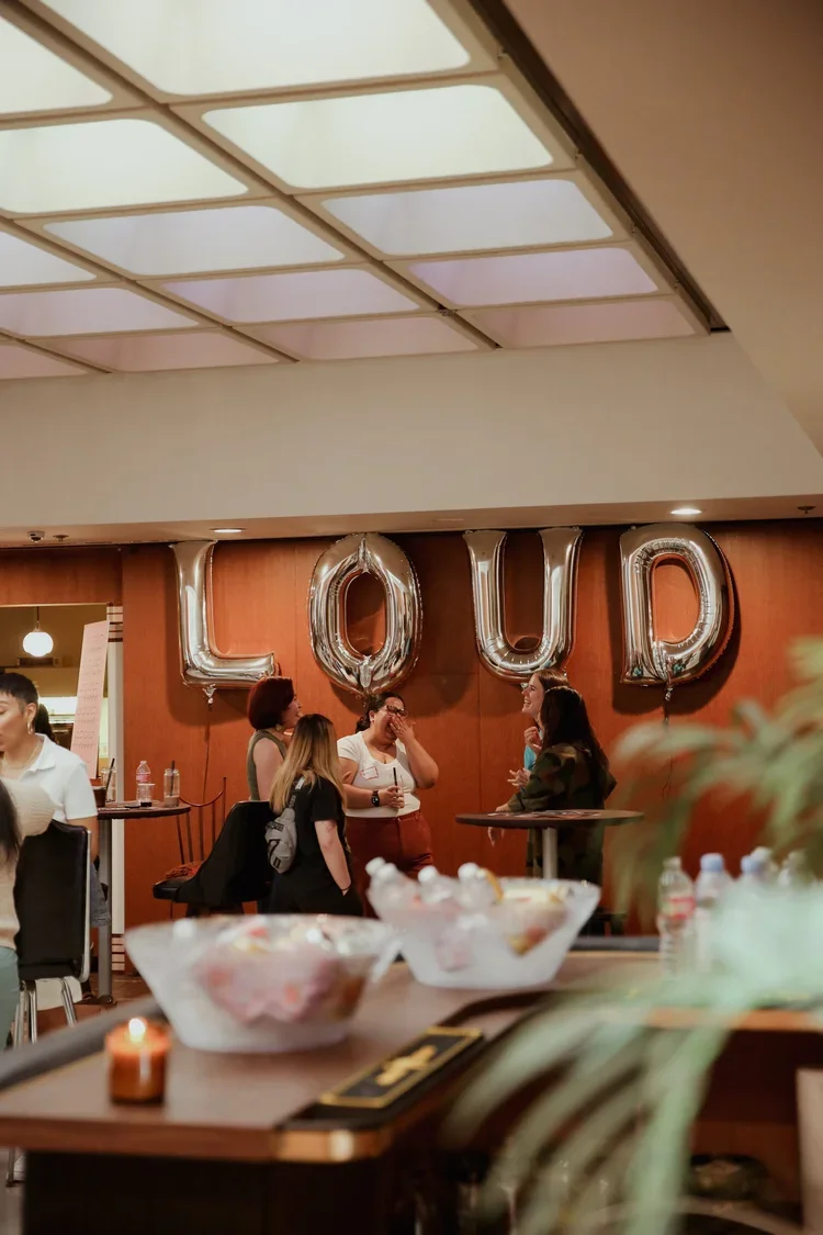 People standing and chatting in a restaurant or cafe with large silver balloons spelling 'LOUP' on the wall behind them