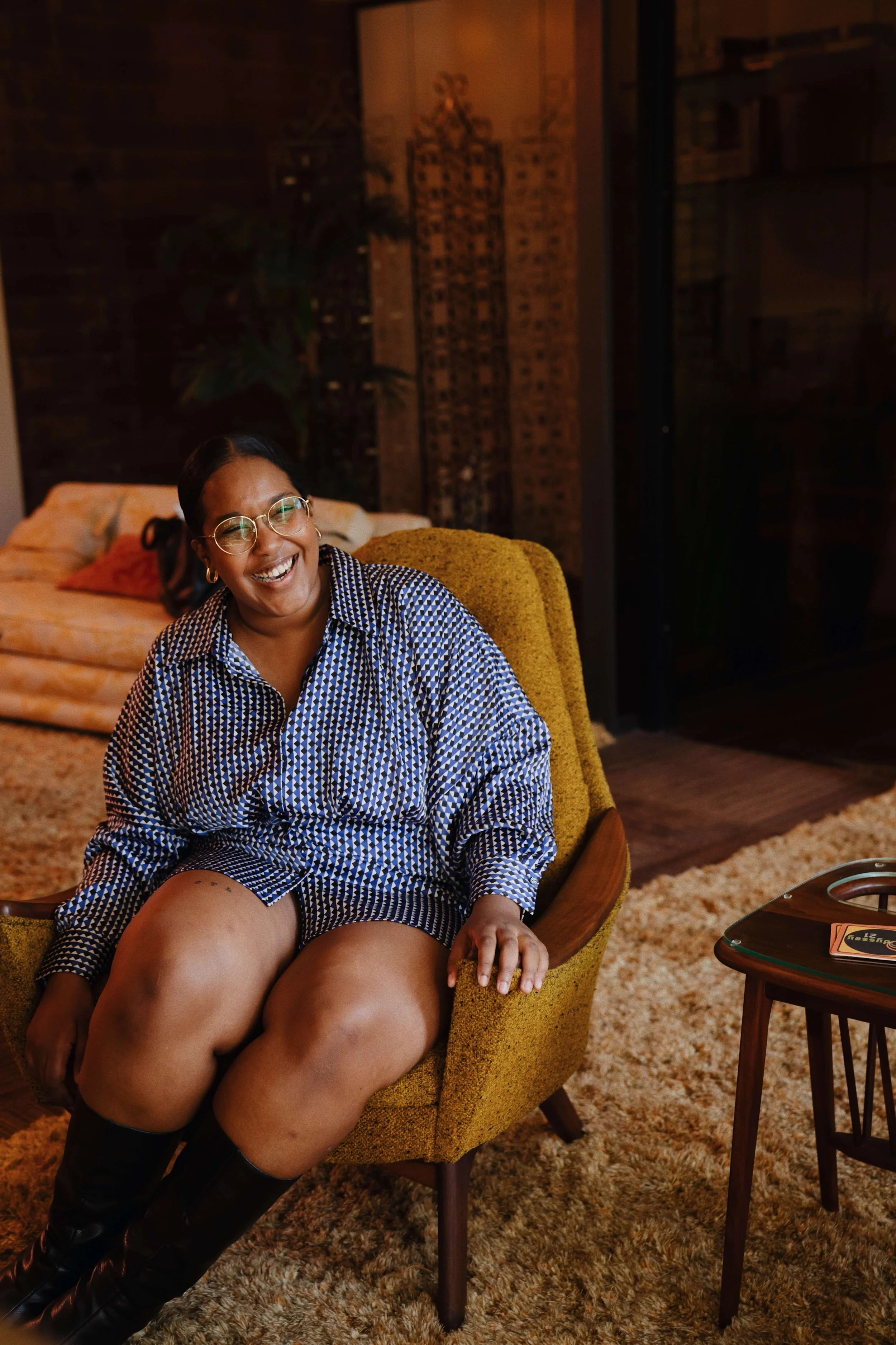 A woman with dark hair and glasses, smiling, is sitting in a yellow armchair. She is wearing a matching patterned shirt and shorts set, and black boots. She is in a room with a vintage-style interior, including a patterned rug, a small round wooden table, a sofa, and a brick wall.