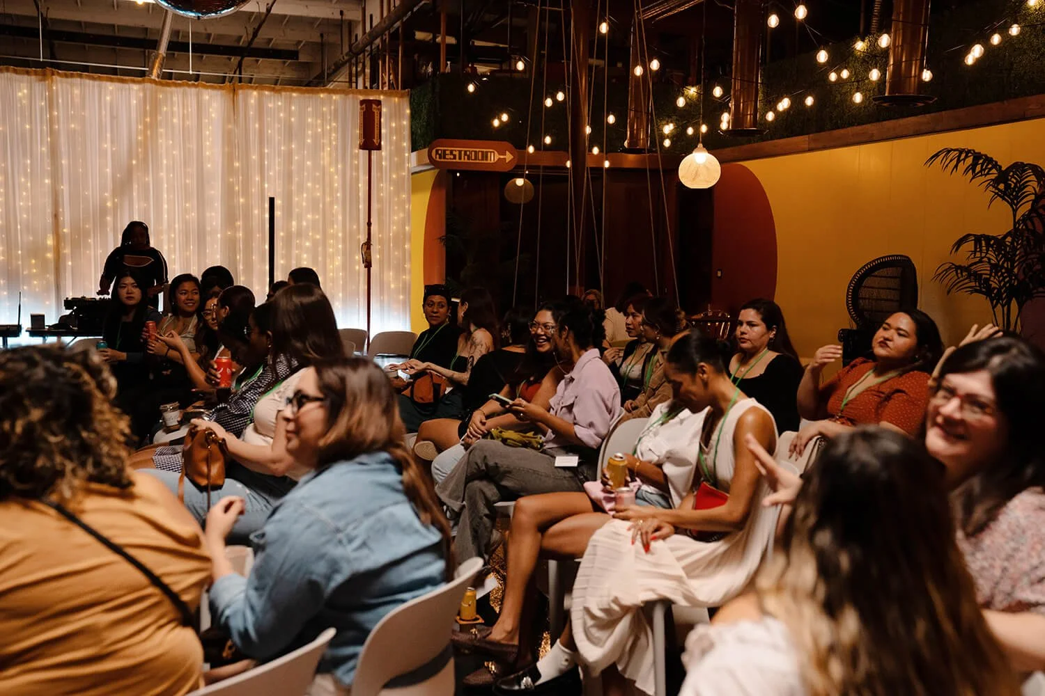 A group of women seated in a dimly lit venue with string lights and sheer curtains, attending an event or presentation.