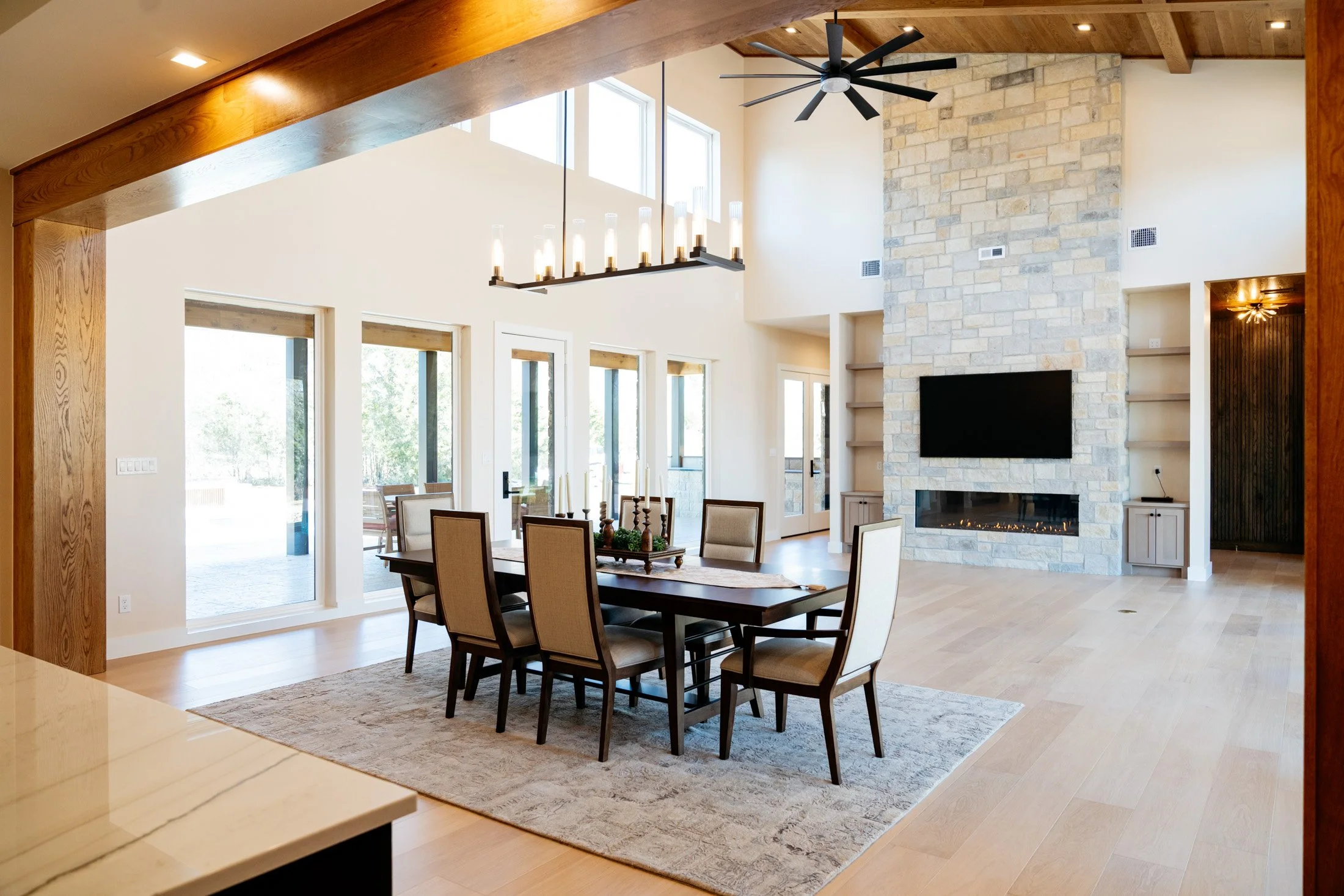 Spacious, modern living room with high vaulted wood ceiling, large windows, stone fireplace with mounted flat-screen TV, and a dining table with chairs. Bright, open, with natural light.