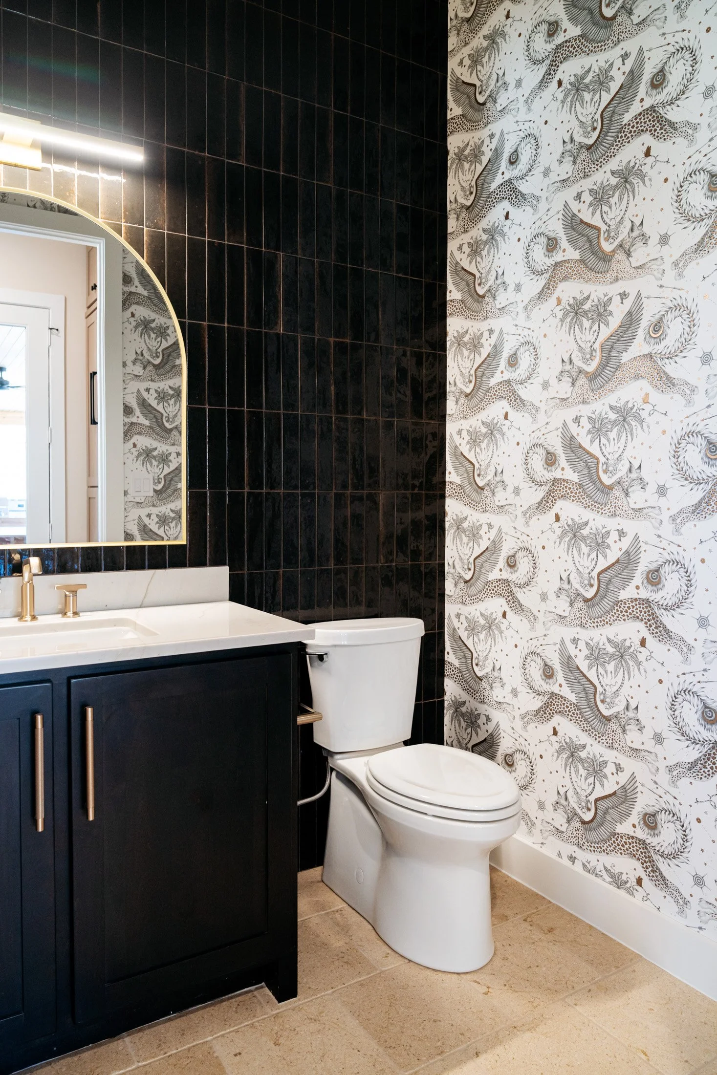 A modern bathroom featuring a black vanity with gold handles, a white countertop with a gold faucet, a large mirror, black tile wall, and a wallpapered wall with white background and leopard and palm tree illustrations.