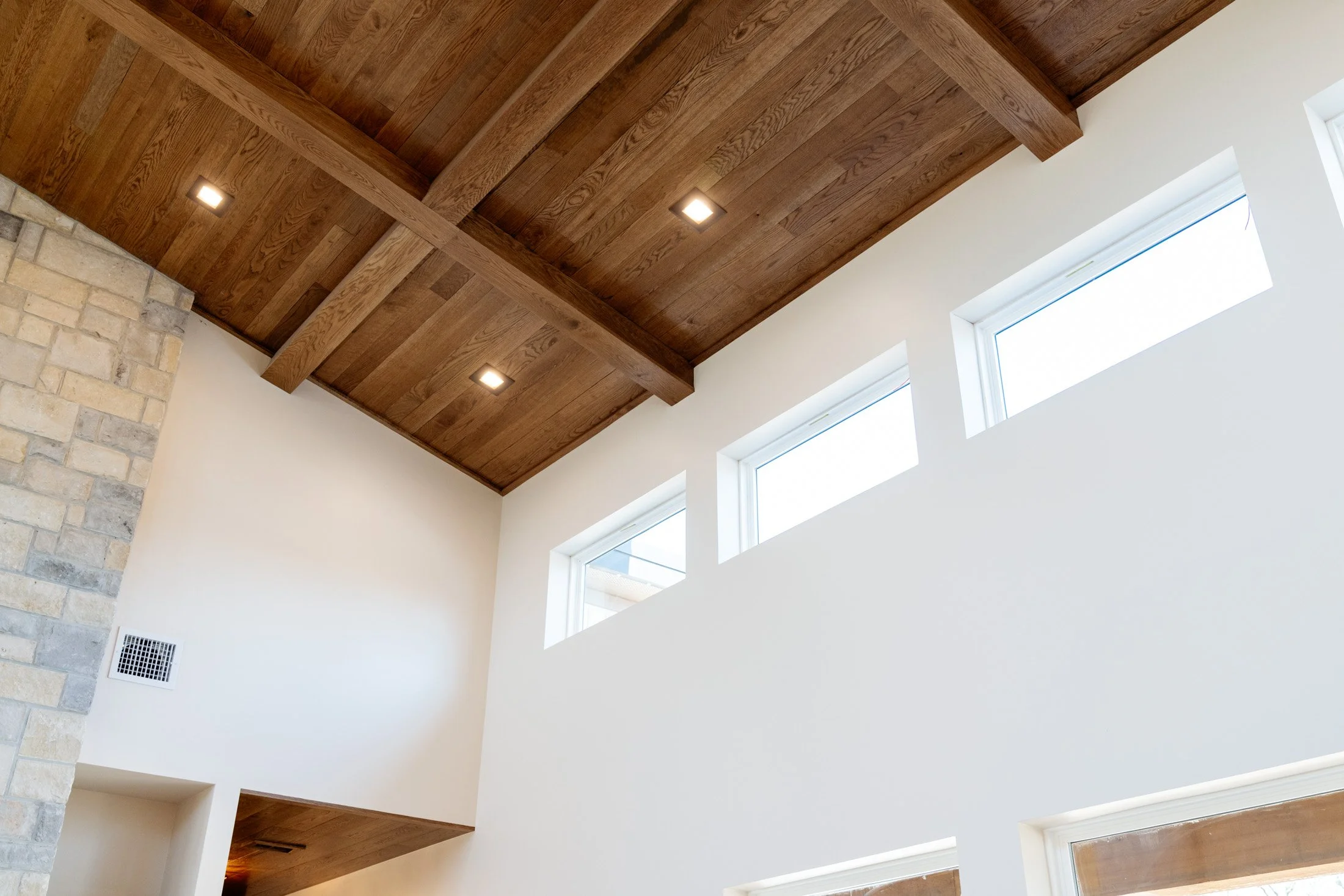 Interior view of a high ceiling with wooden paneling, three rectangular ceiling lights, a stone wall on the left, and large windows on the right letting in natural light.