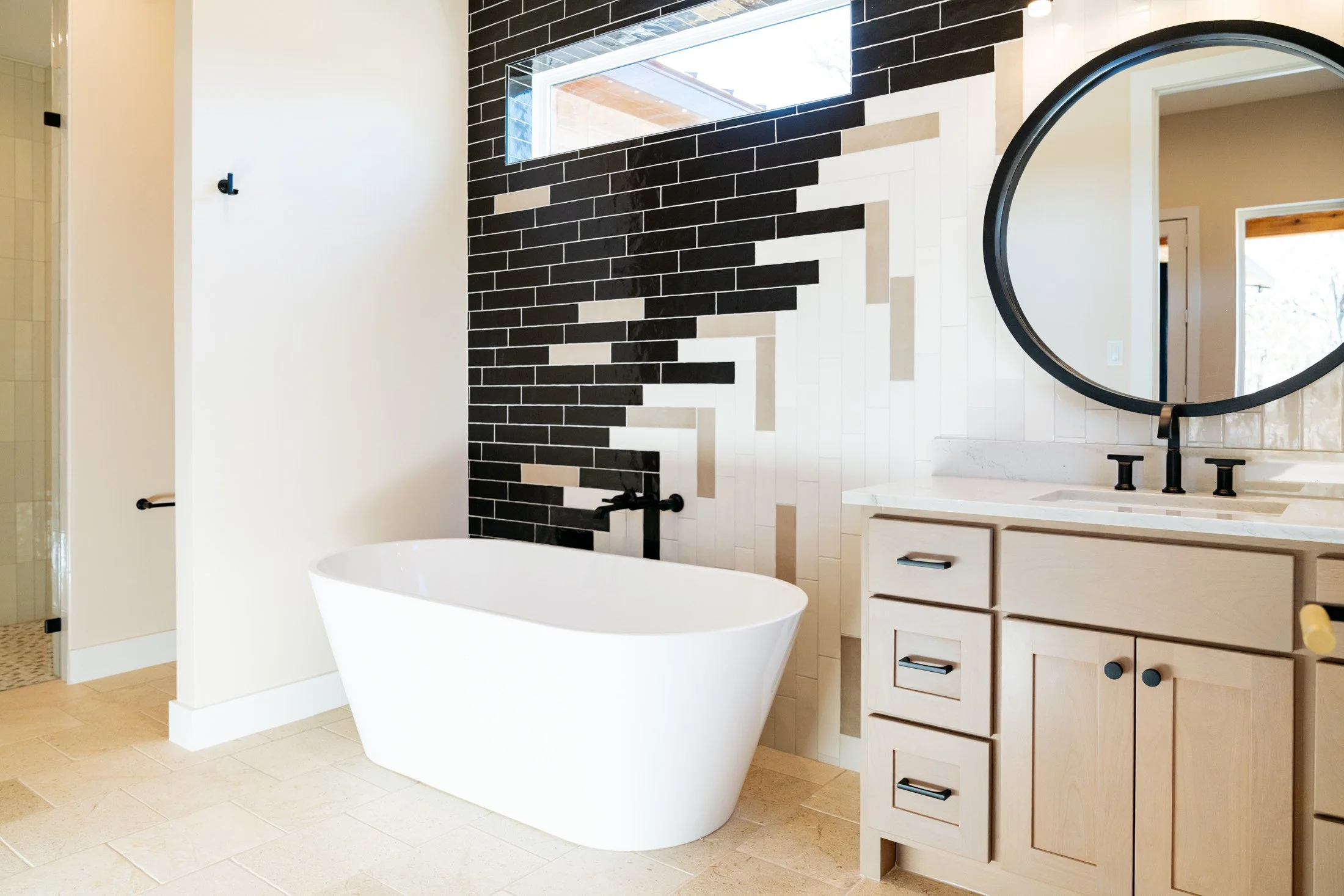 Modern bathroom with a white freestanding bathtub, black and beige tiled accent wall, wooden vanity with drawers, and a round black-framed mirror.