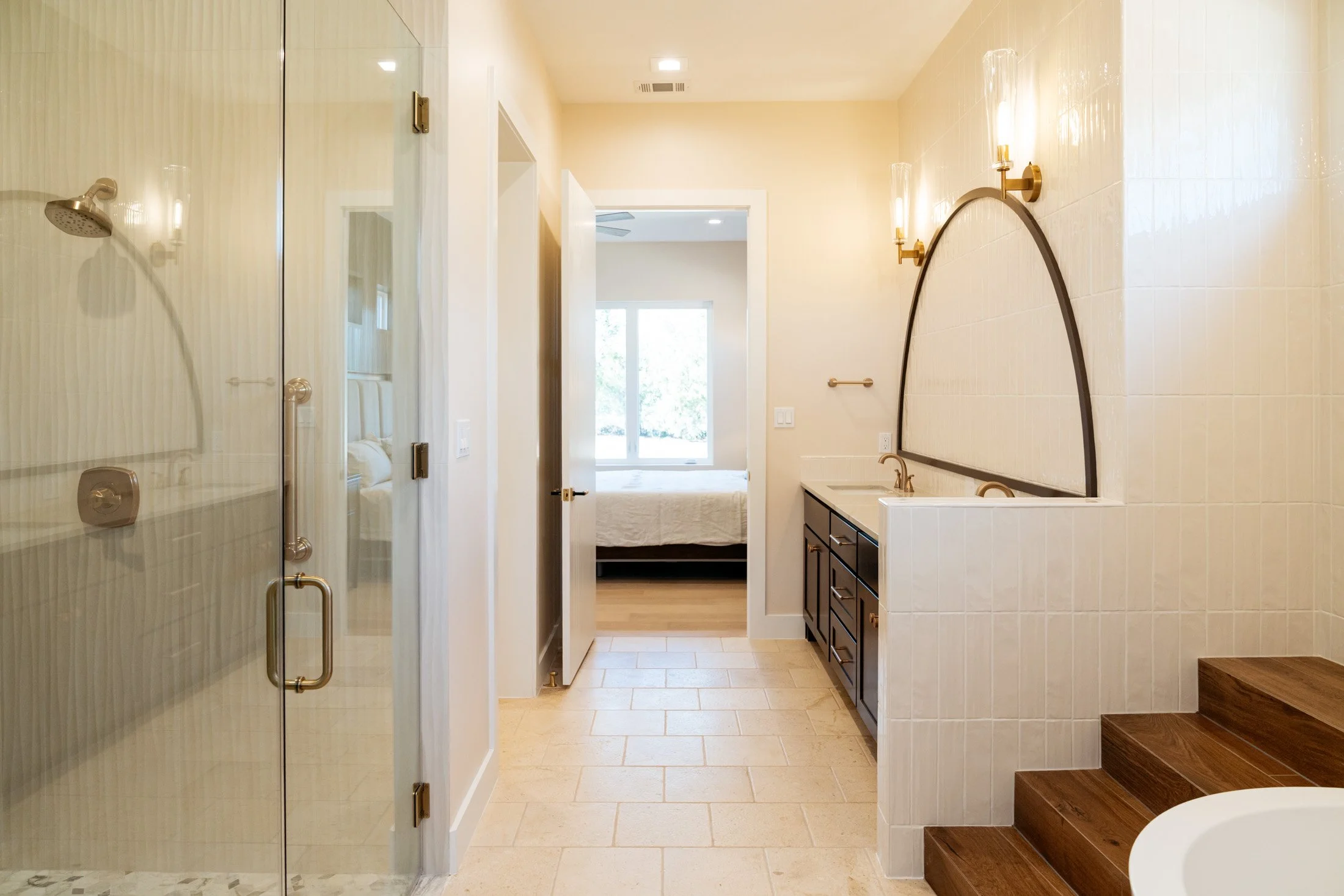 A spacious bathroom with a glass shower on the left, a vanity with dark cabinets and a white countertop on the right, and a mirror with wall sconces above. There is a doorway leading to a bedroom with a bed, large window, and ceiling fan in the backg